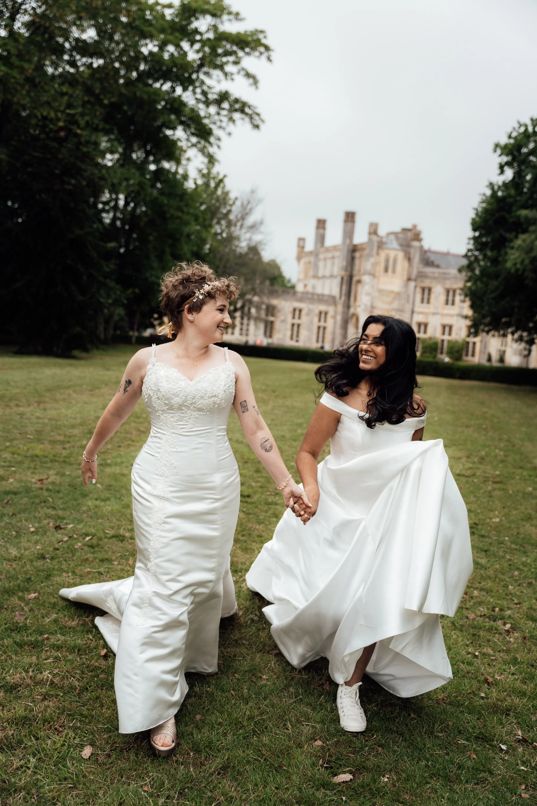 Two brides in wedding dresses are holding hands and walking across a grassy field with a castle in the background.
