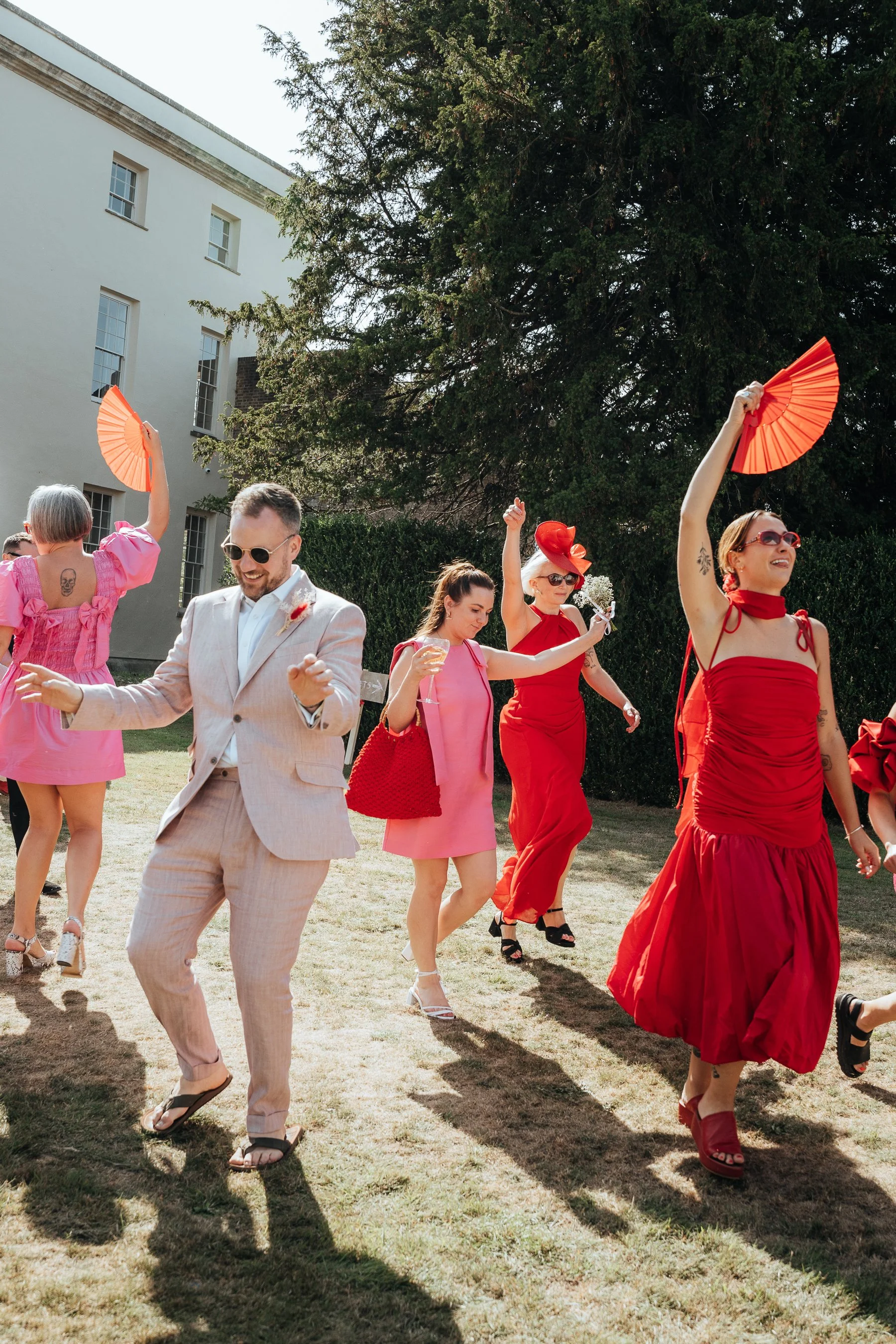 People dancing at an outdoor wedding, dressed in colourful clothing, holding fans, with a white building and tall trees in the background.