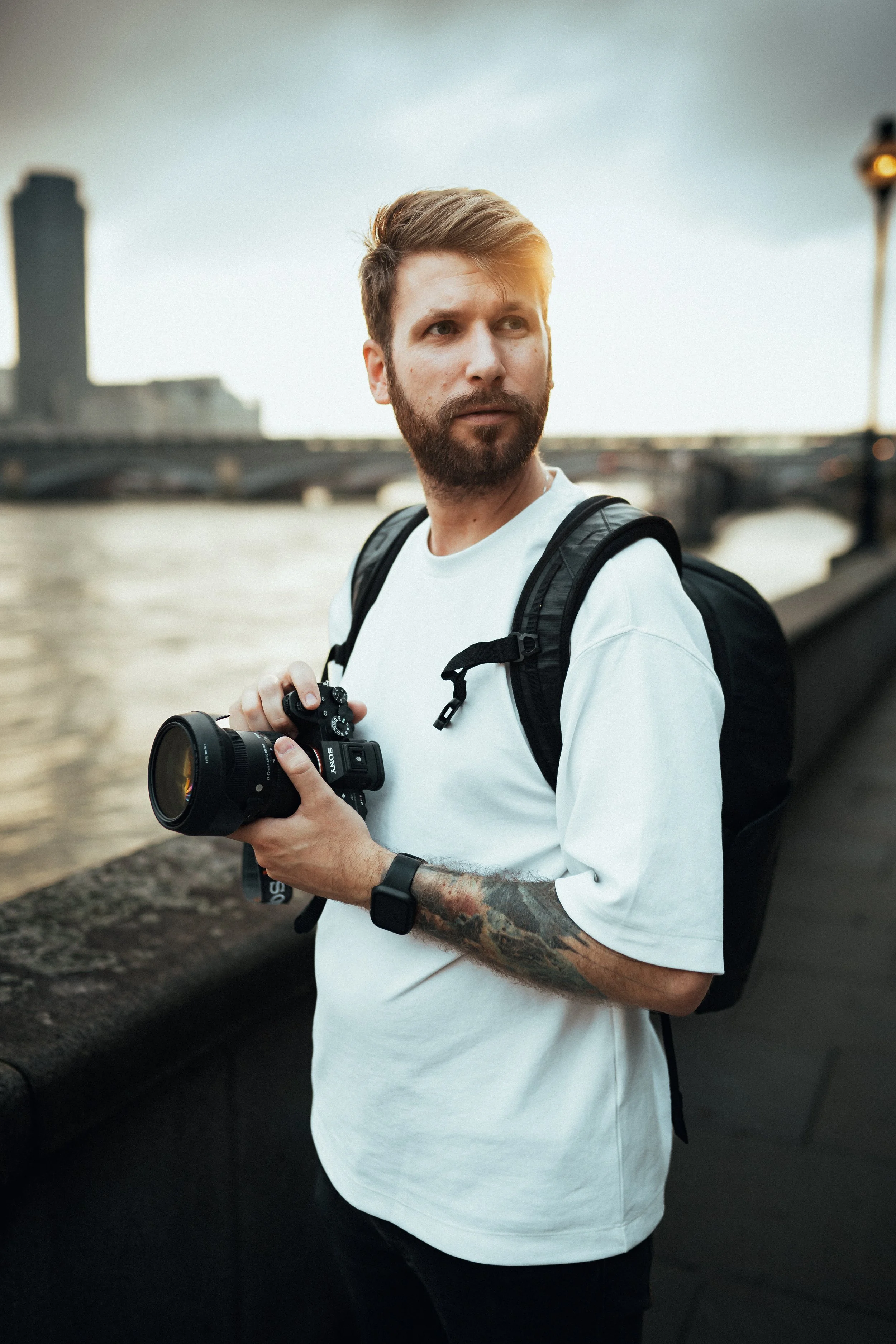 A man with a beard and tattoos on his left arm holding a camera, standing near a river with a bridge and city buildings in the background.