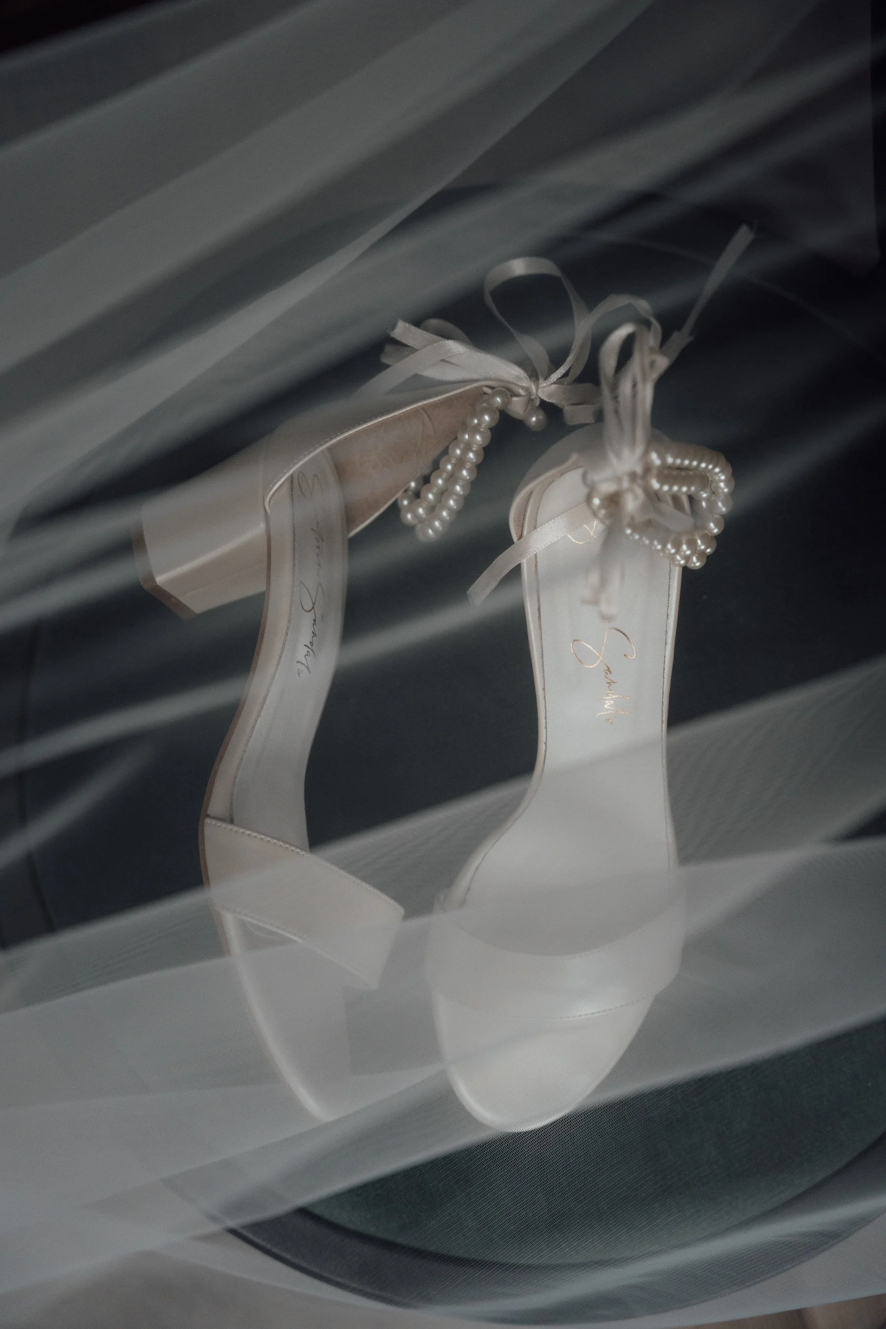 White high-heeled shoes with ribbons and pearl detail are hanging inside a closet with shelves and a gray surface.