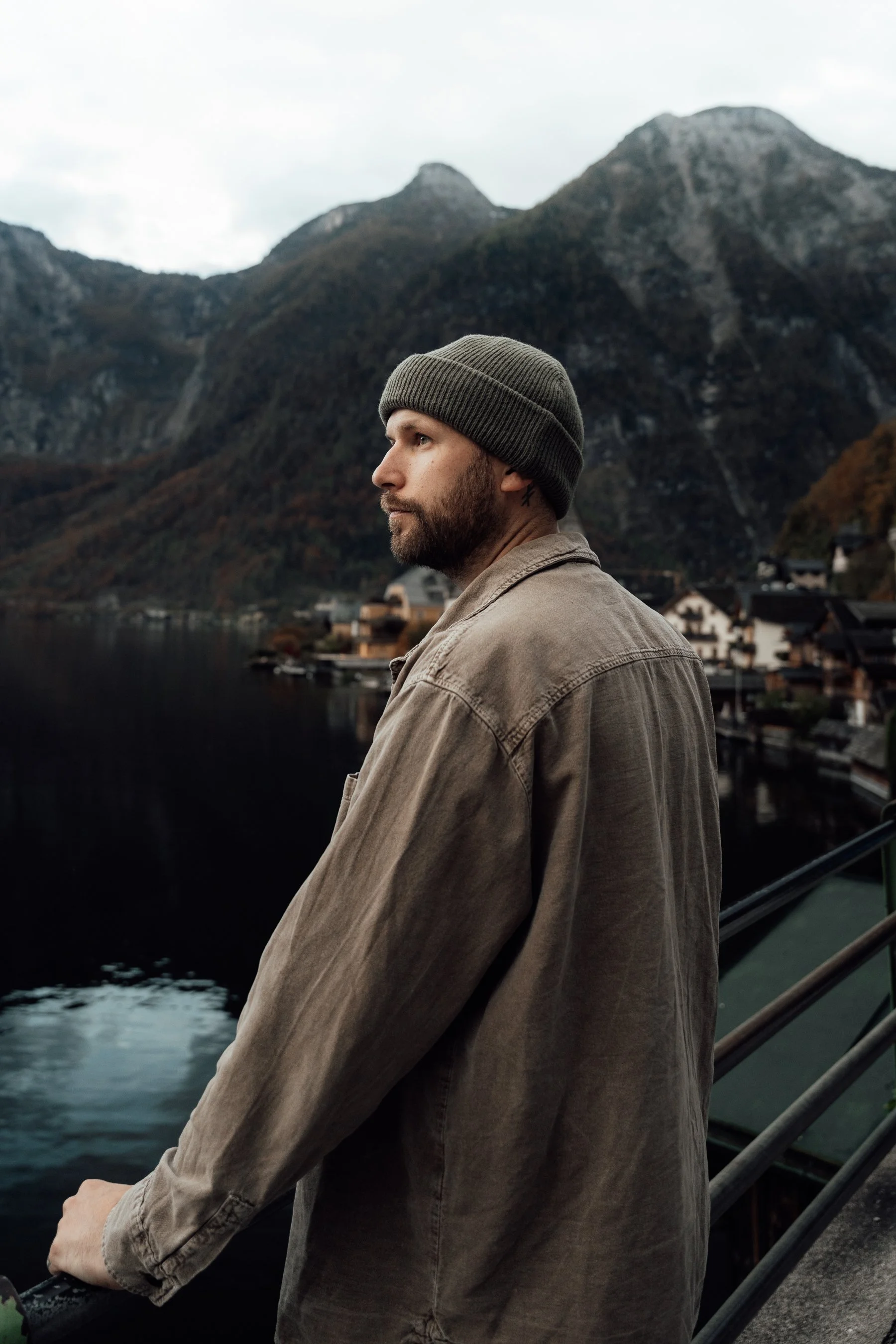 A man with a beard and knit cap standing on a bridge overlooking a lake and mountains in the background.