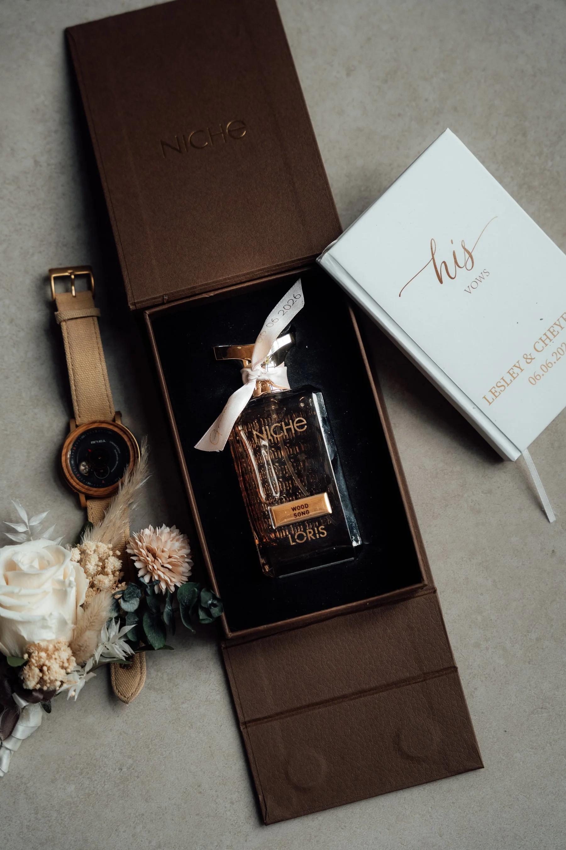 A gift set including a perfume bottle labeled 'Niche', a bouquet of white and beige flowers, a beige watch, and a white wedding or special occasion program with gold lettering, all arranged on a neutral surface.