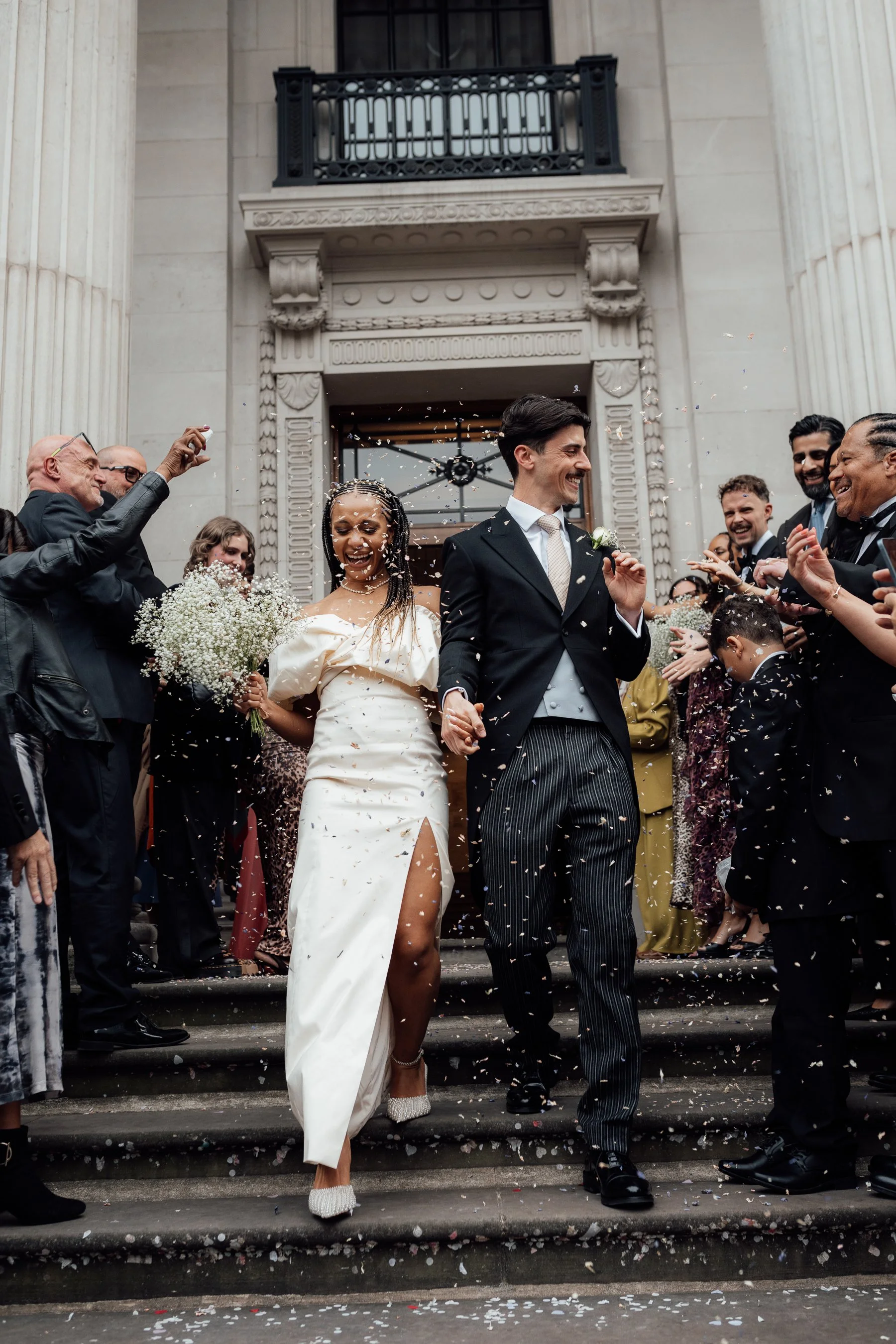 A newly married couple walking down the stairs, surrounded by friends and family throwing confetti, celebrating outside a historic building with large columns and ornate architecture.