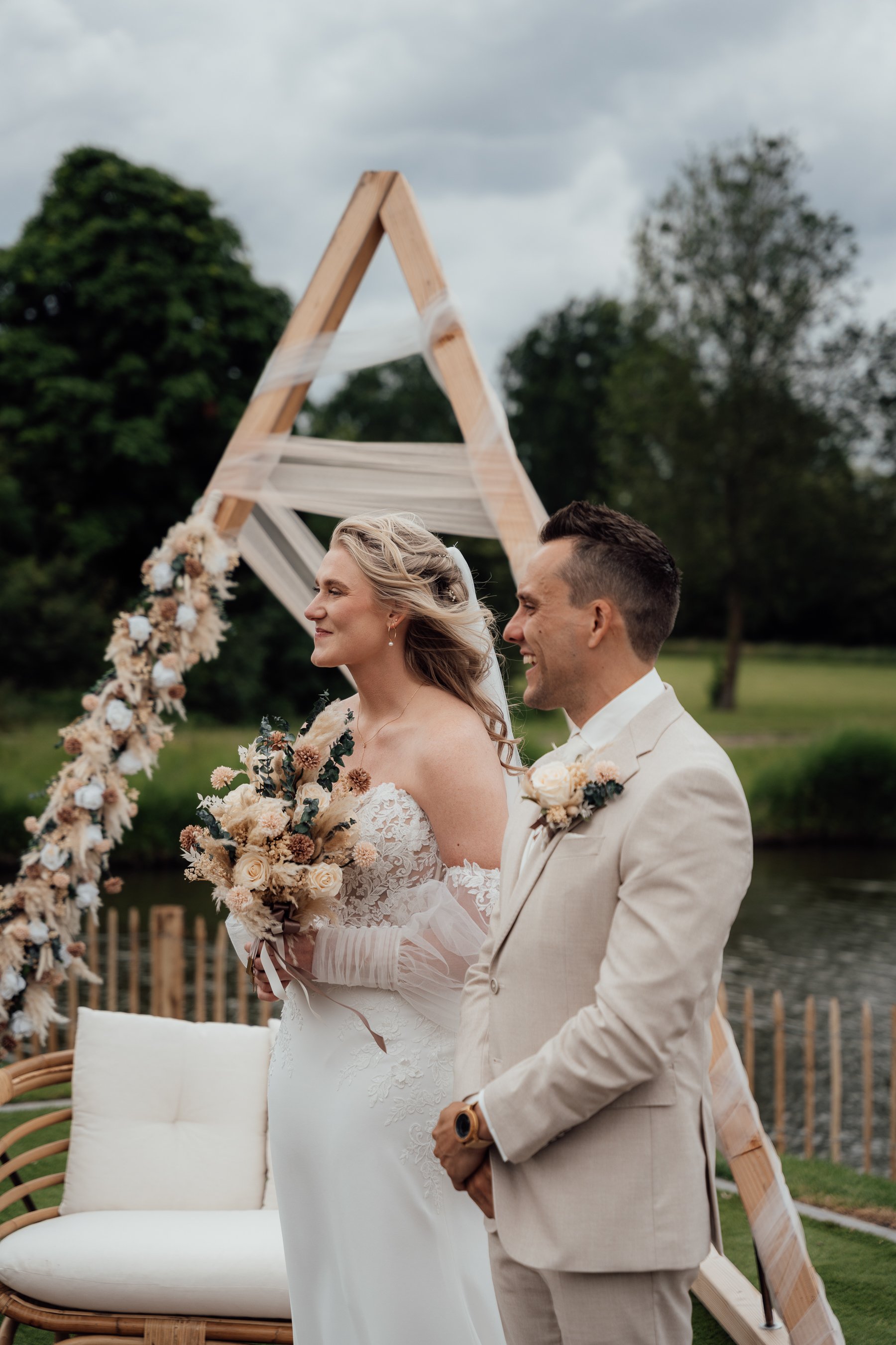 A bride and groom standing outdoors during their wedding ceremony, with a decorative wooden arch in the background and greenery surrounding them.