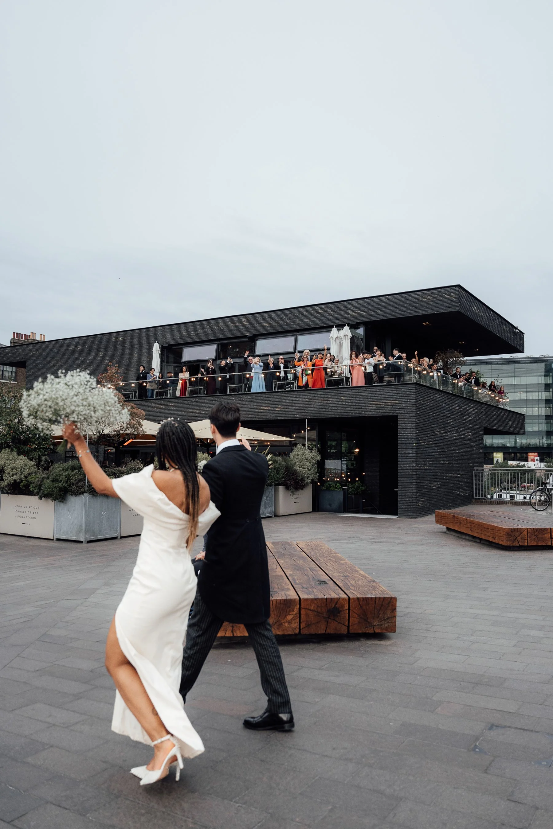 Couple dancing in formal attire outdoors, with a group of people on the balcony above them celebrating, at a modern building with dark brick exterior under cloudy sky.