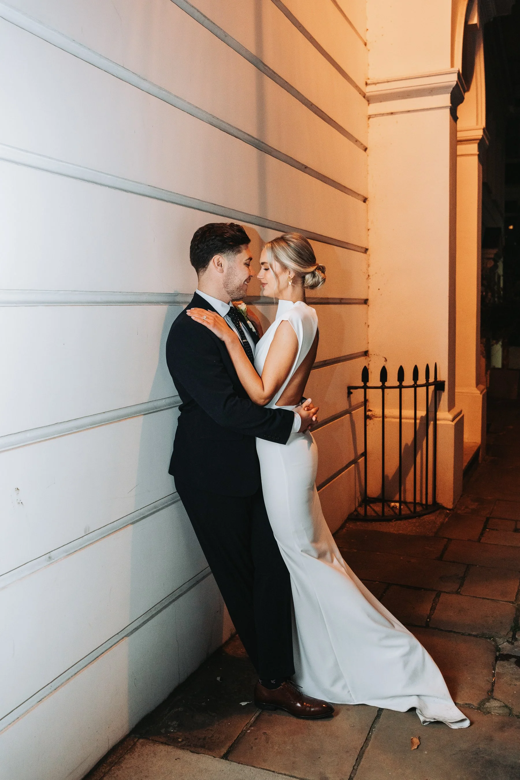 A bride and groom standing close together, touching foreheads with eyes closed, against a white wall outside at night, both smiling softly. The groom wears a black tuxedo, and the bride wears a white gown with a low back, her hair styled in a low bun