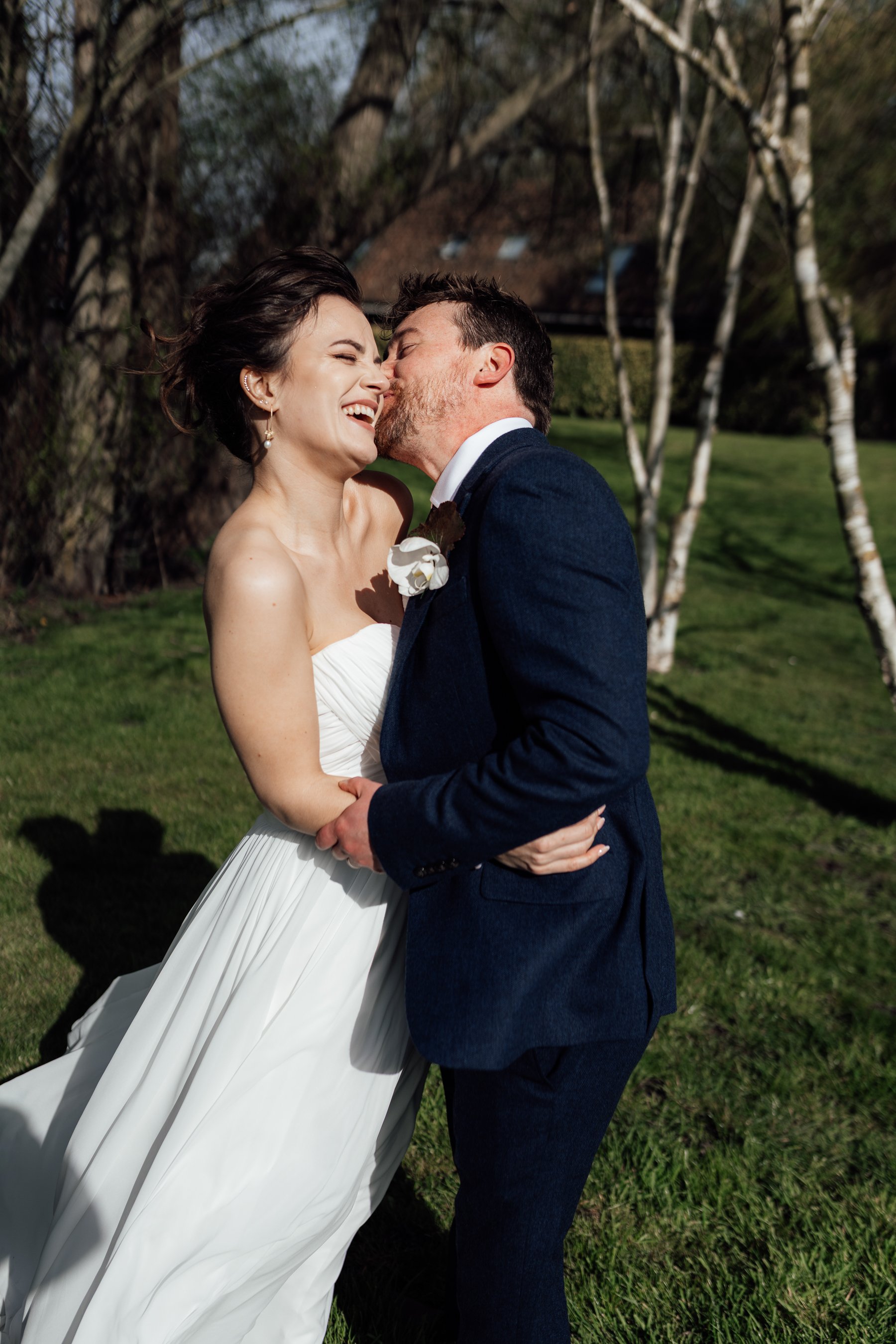 A bride and groom sharing a kiss outdoors during daytime, with trees and grass in the background.