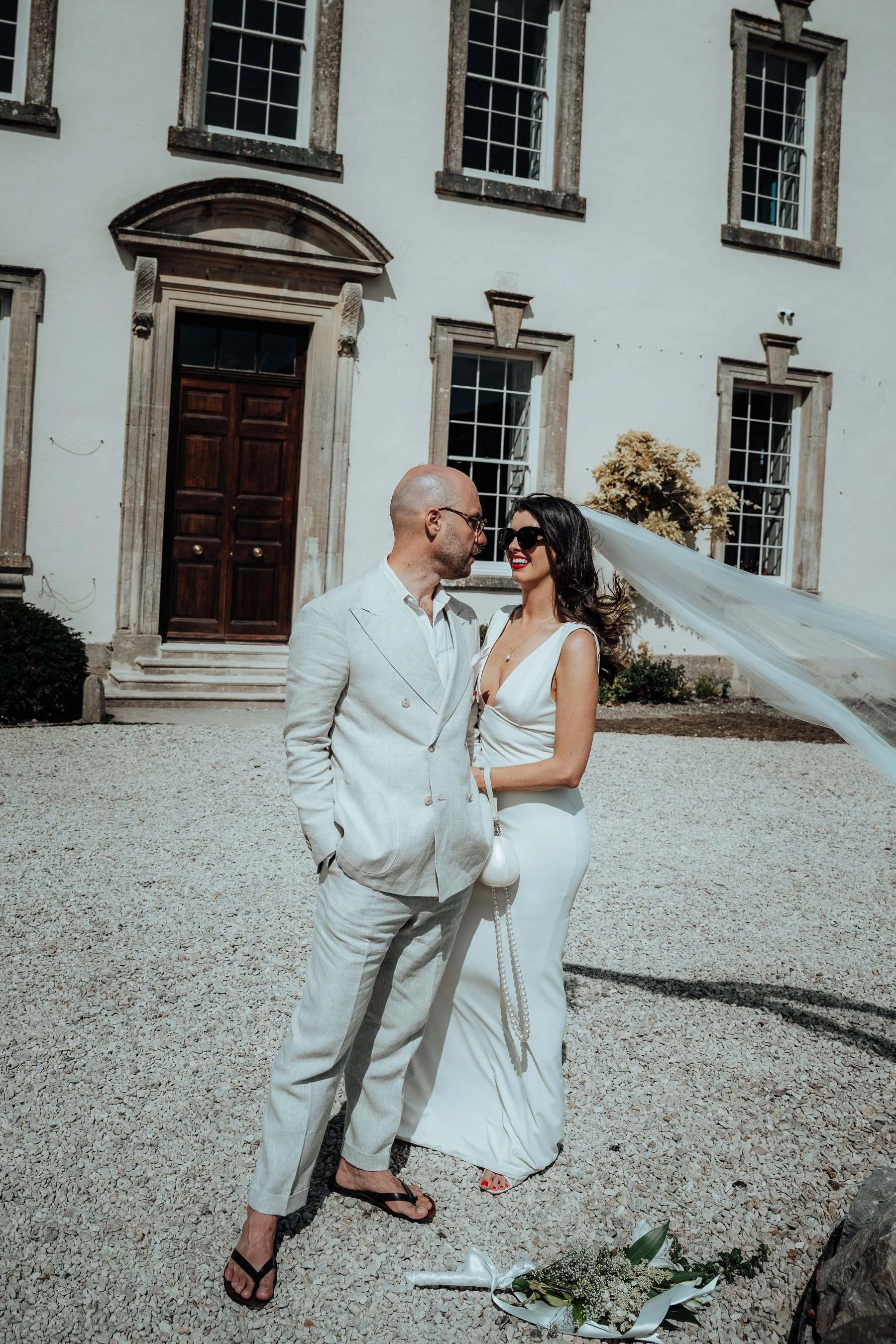 A newly married couple standing outside a historic building, dressed in wedding attire, smiling and looking at each other. The man is in a light-colored suit with sandals, and the woman is in a white dress with a veil, holding a bouquet of flowers pl