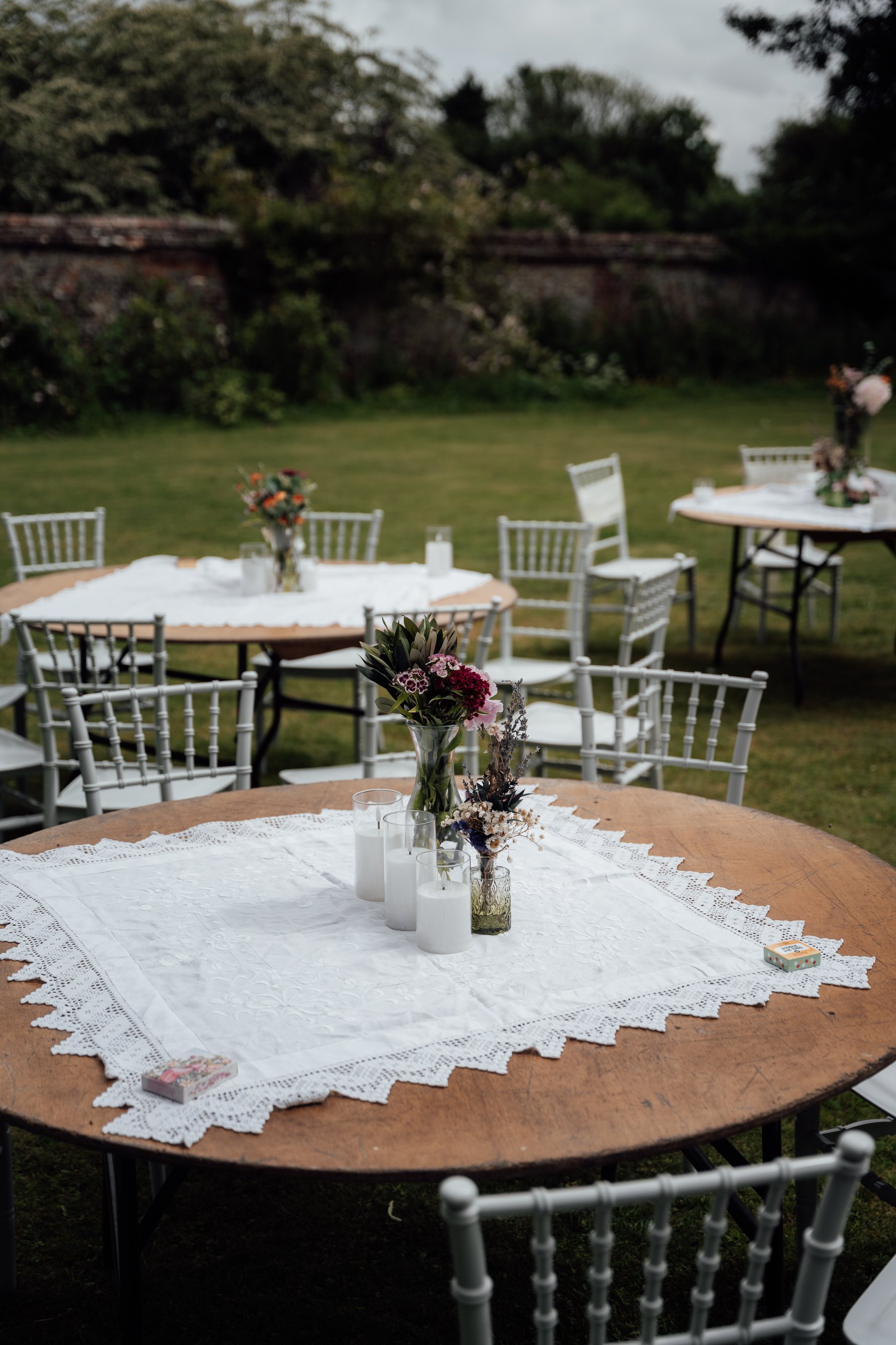 A round wooden outdoor table with a white lace tablecloth, decorated with a clear vase of mixed flowers, white candles, and a box of matches, set in a grassy yard with other similar tables and white chairs.