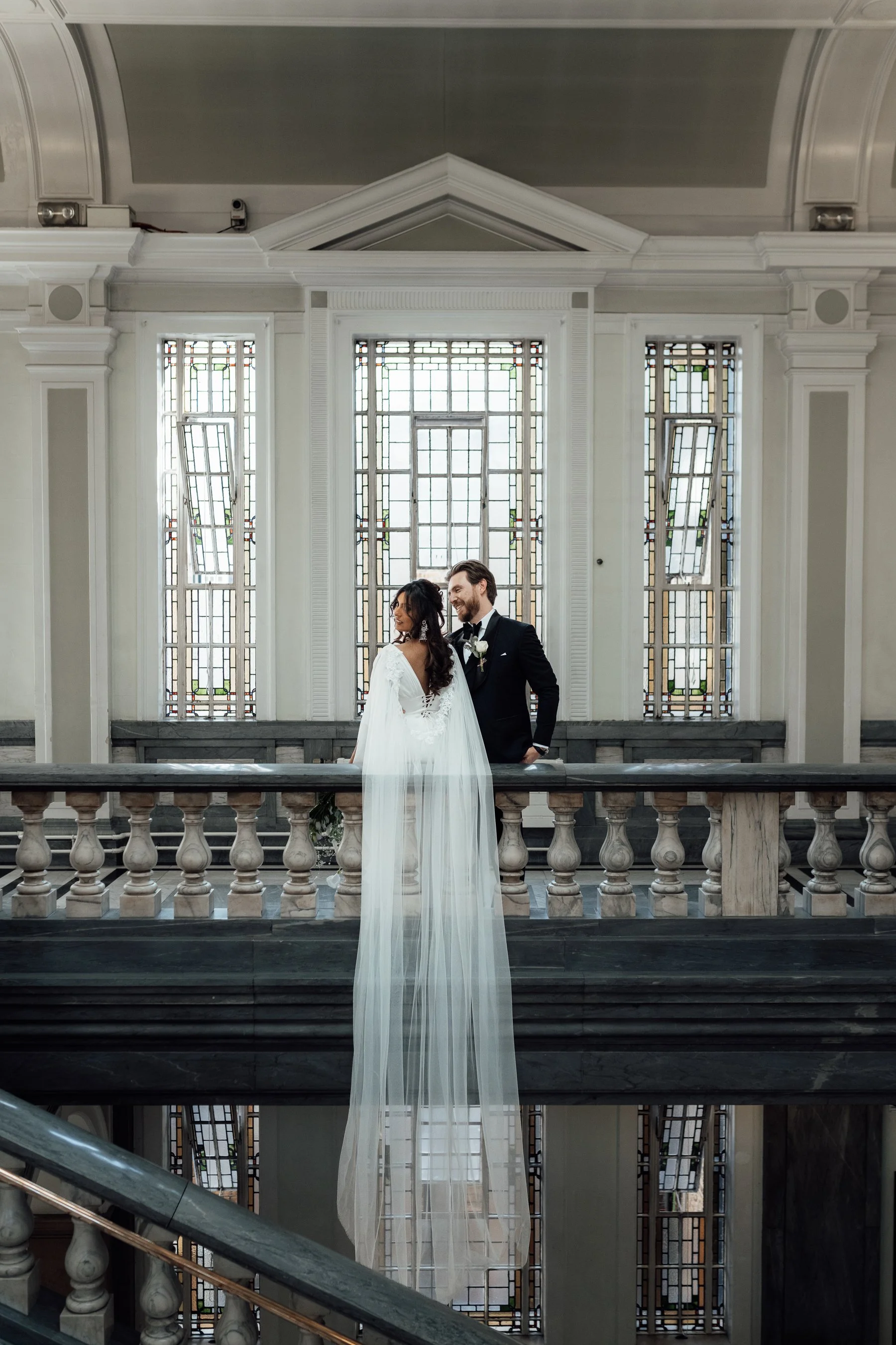 A bride and groom standing on a marble staircase in front of stained glass windows inside a church, smiling and facing each other.