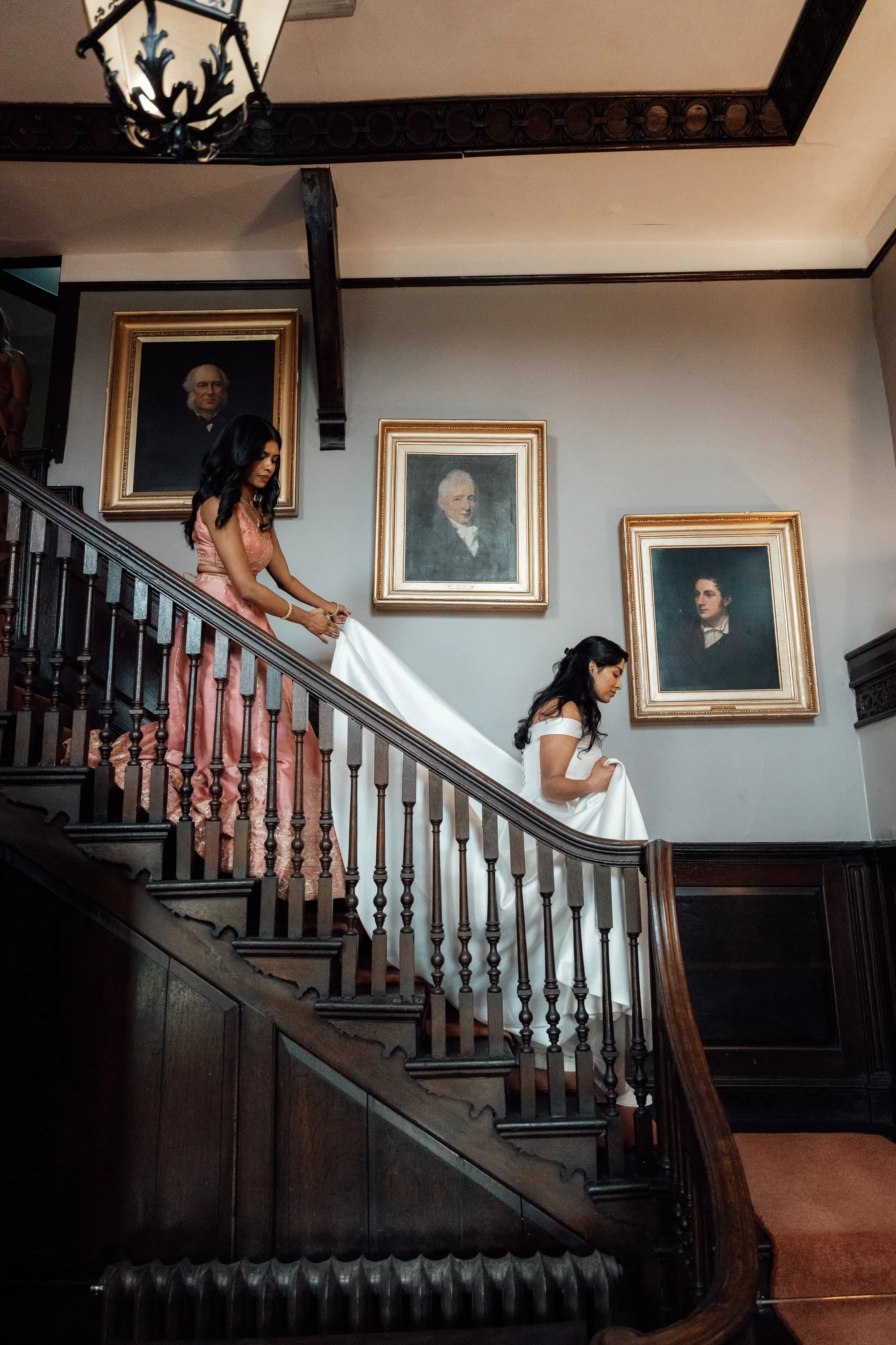 A bride in a wedding dress descending a dark wooden staircase in a room with framed portraits on the wall.