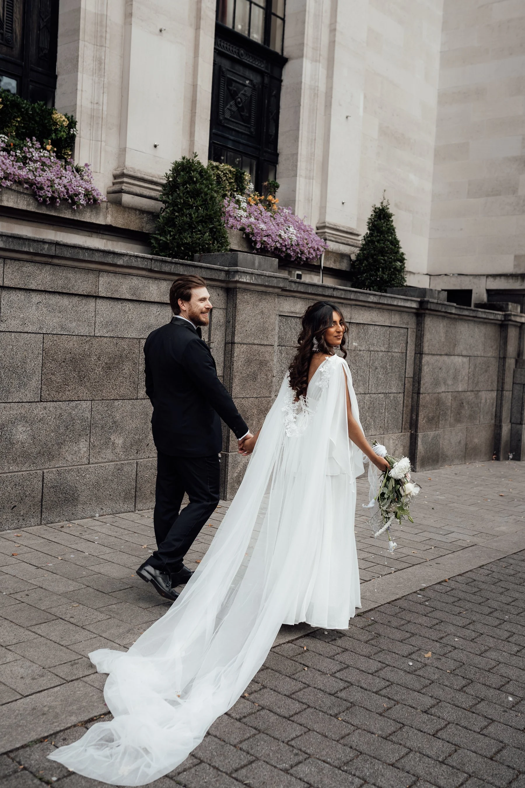 A bride and groom holding hands walking outside a building with stone walls, flowers, and potted bushes.