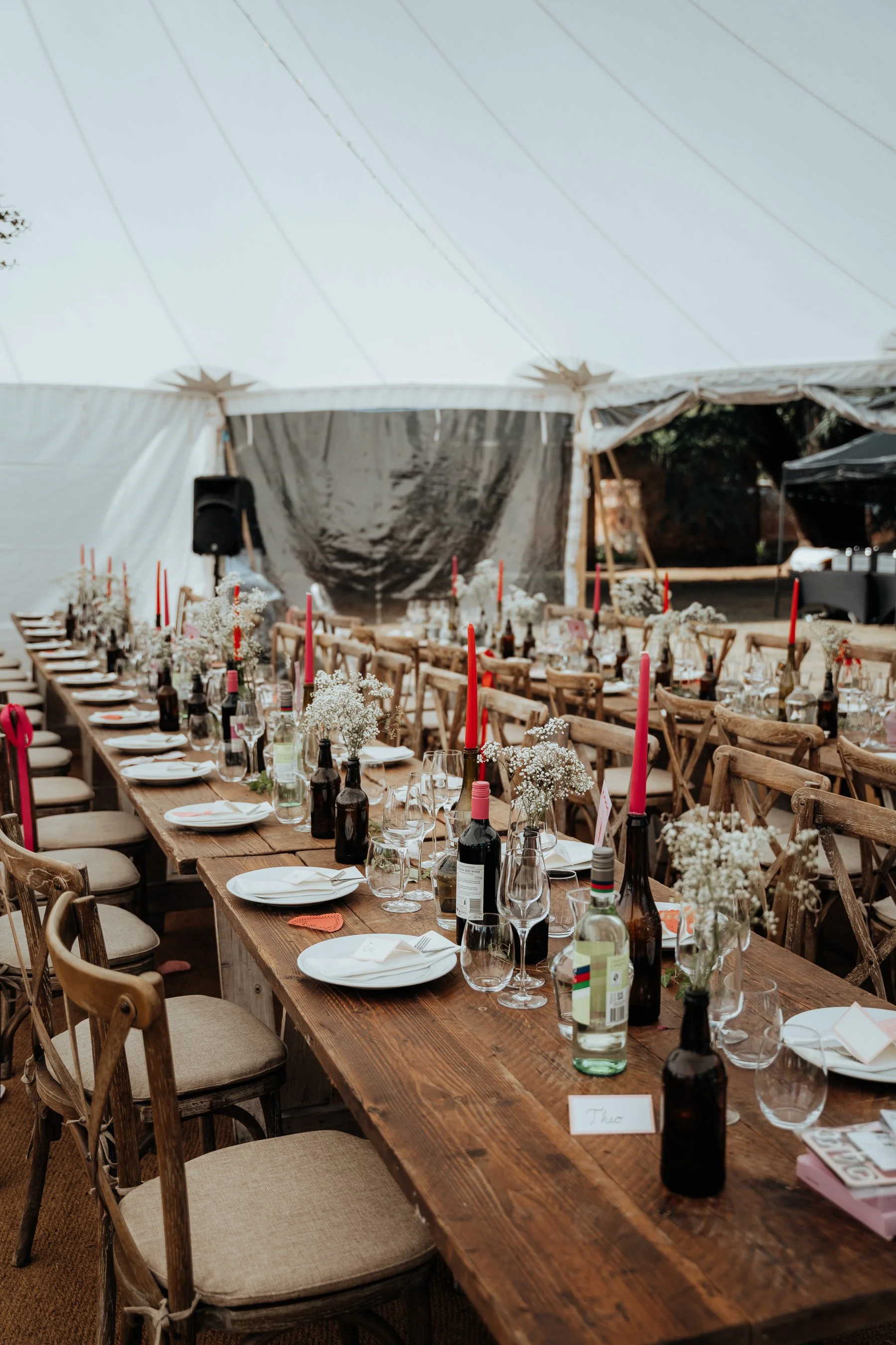 Long wooden dining table set for a special event with wine bottles, glasses, plates, flowers, and pink candles, under a large white tent with open sides.