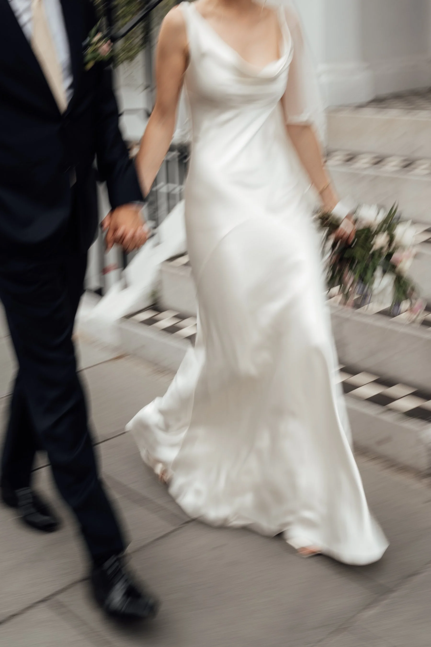 Blurred image of a bride wearing a white wedding dress and a groom dressed in a black suit holding hands while walking outdoors on a sidewalk.