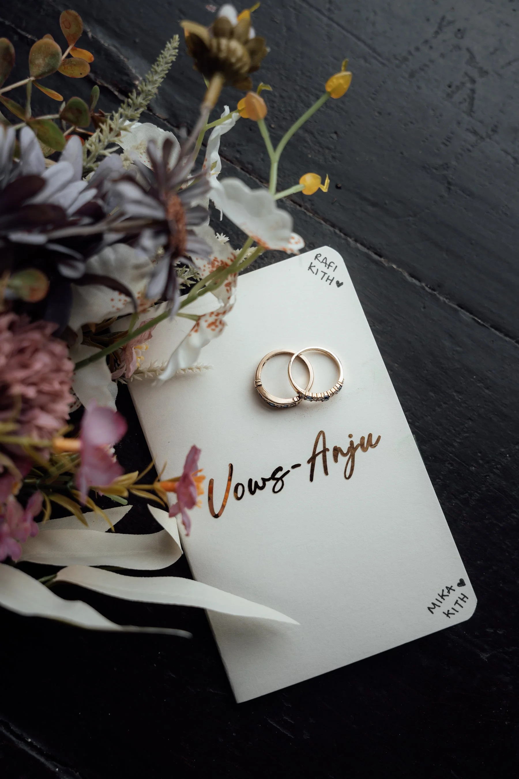 A white card with handwritten text reading 'Vows to Annie' and two wedding rings placed on top. Surrounding the card are various artificial flowers in pink, purple, green, and white, with a small note reading 'Mika Kith' in a heart shape. The backgro