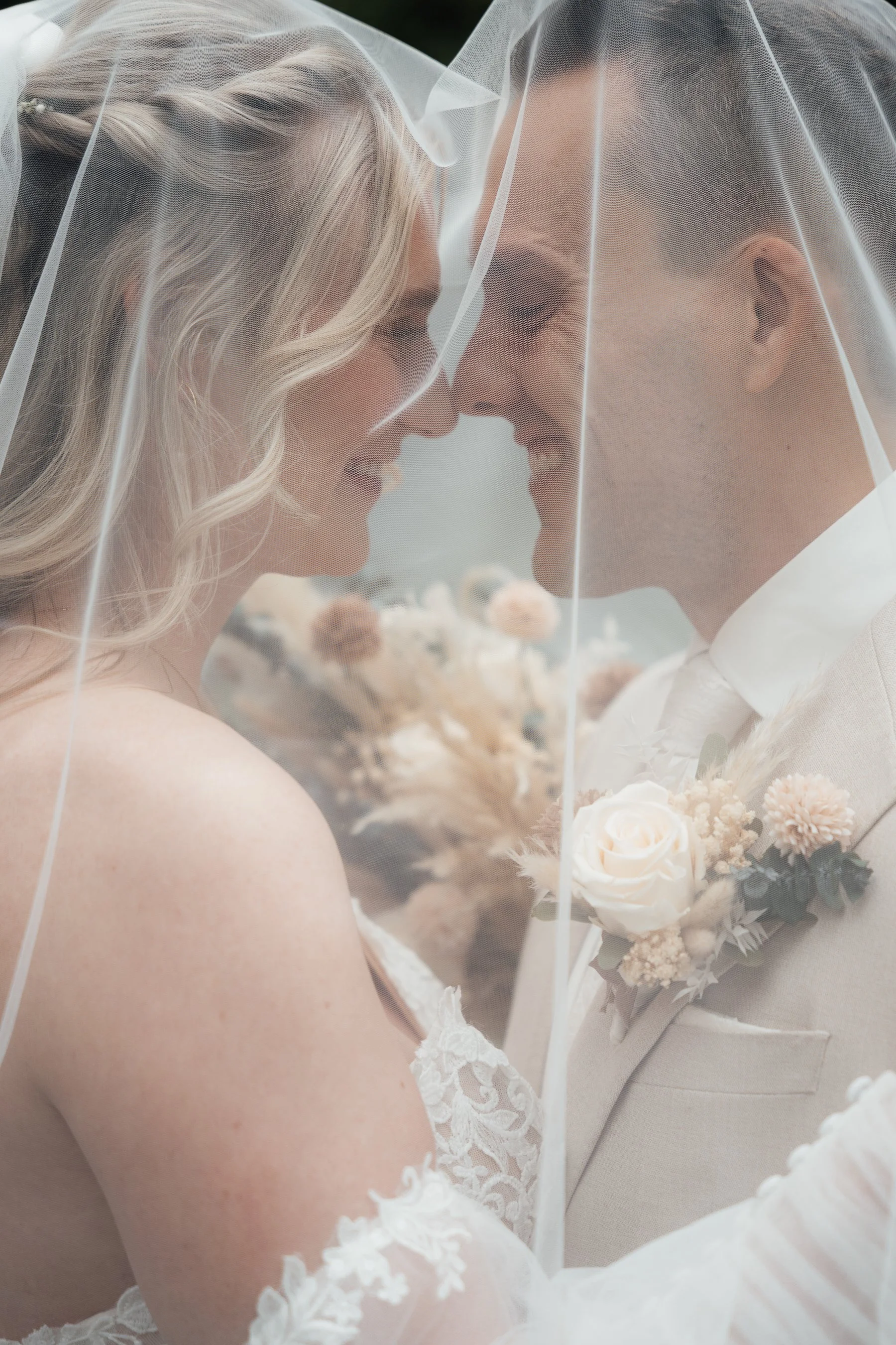 Close-up of a bride and groom with foreheads touching, under a veil, holding a bouquet of white and blush flowers, with lace details on the bride's dress.