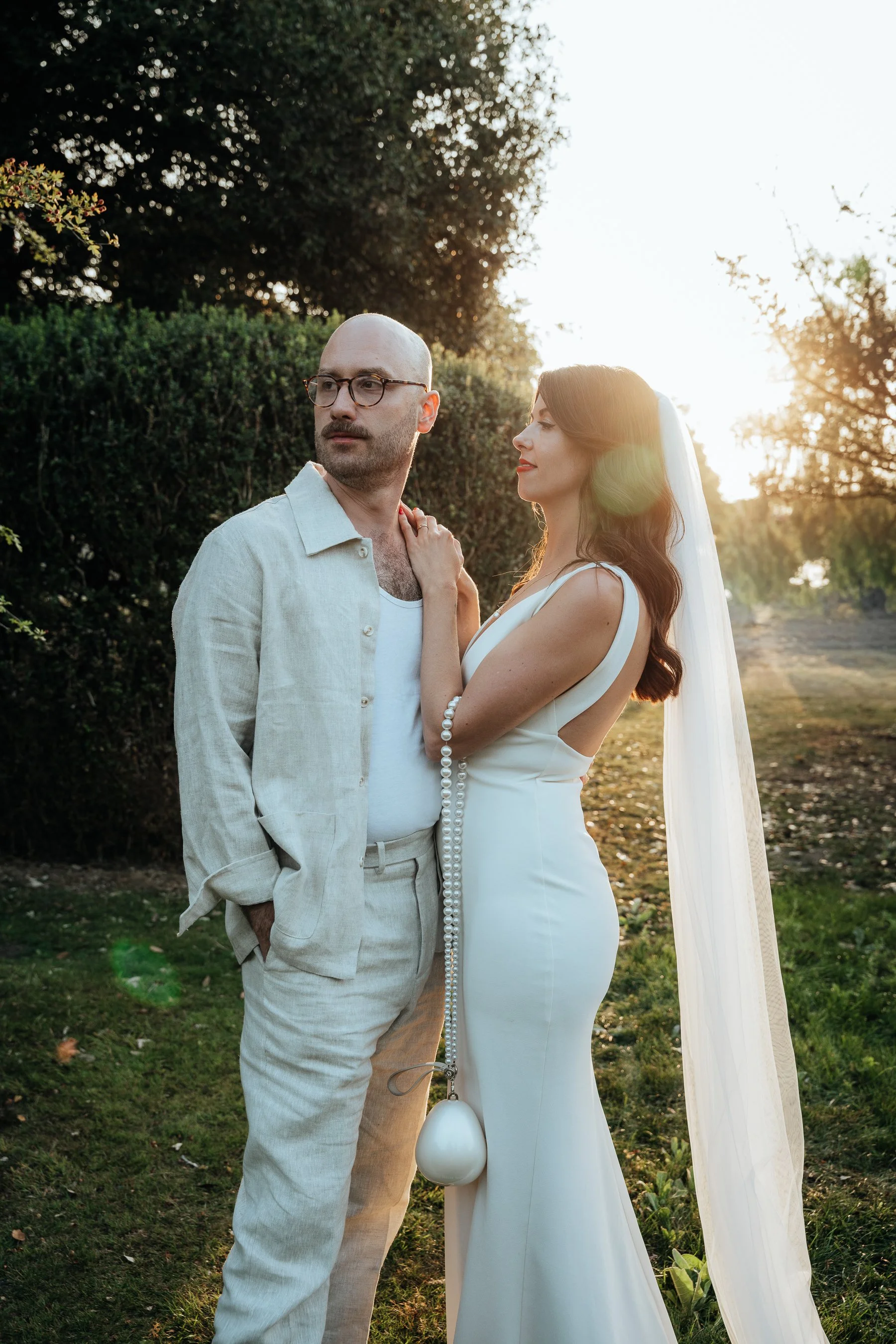 A couple dressed in wedding attire, standing outdoors during sunset with trees and greenery in the background.