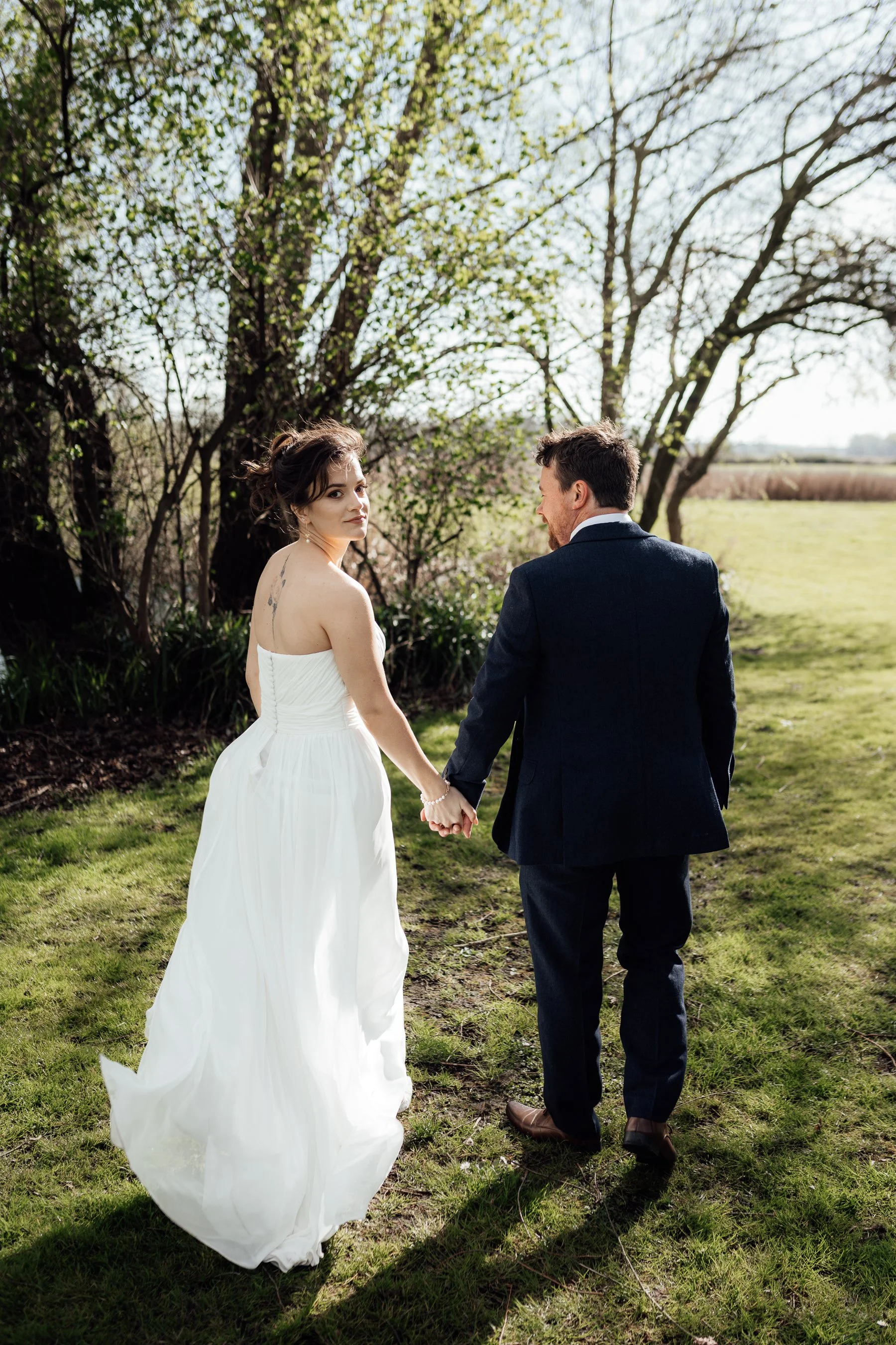 A bride and groom holding hands and walking outdoors on a grassy area with trees and open fields in the background during daytime.