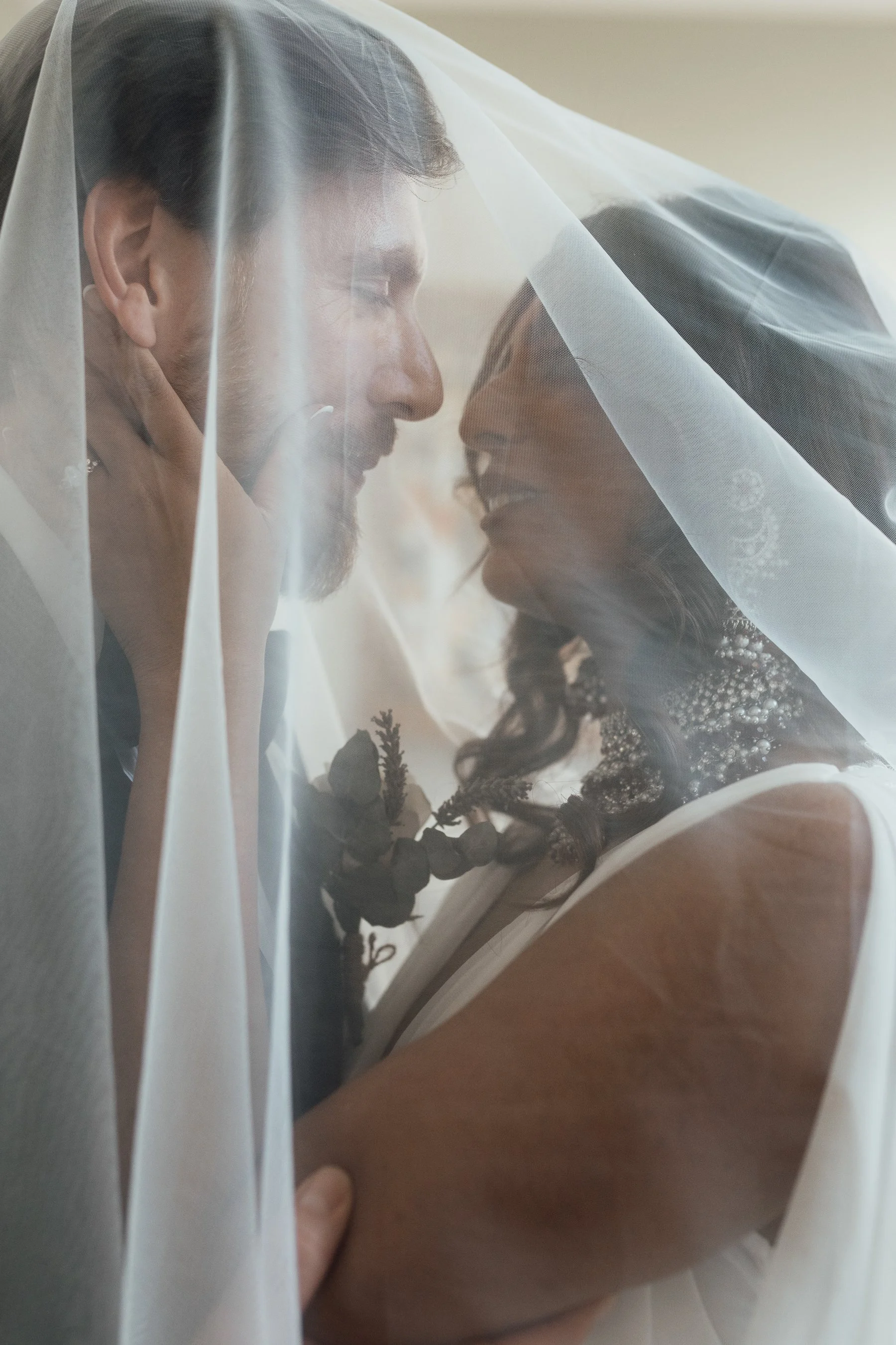 A close-up of a couple behind a sheer white veil, smiling and tenderly touching each other's faces during a wedding or special moment.