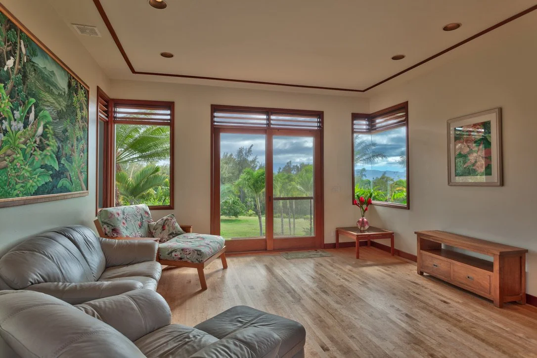 Living room with wooden floors, white walls, a leather sofa, a floral armchair, a side table with flowers, large windows showing a lush green landscape, and framed artwork on the walls.