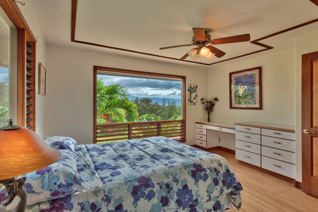 A bedroom with a large window showing a scenic view of trees and mountains, a bed with a floral bedspread, white cabinets against the wall, and a ceiling fan.