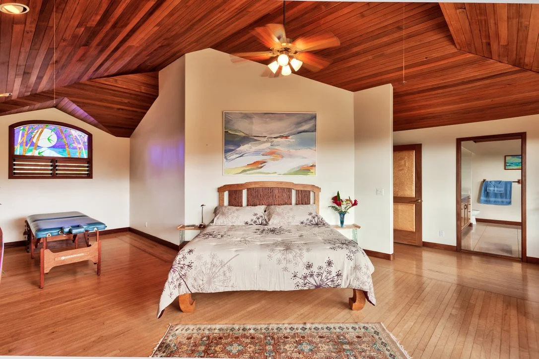 A bedroom with wooden ceiling and floors, a bed with floral bedding, an abstract painting above, a ceiling fan, a small table with flowers, a bench near a stained glass window, and a doorway leading to a bathroom with blue towels.
