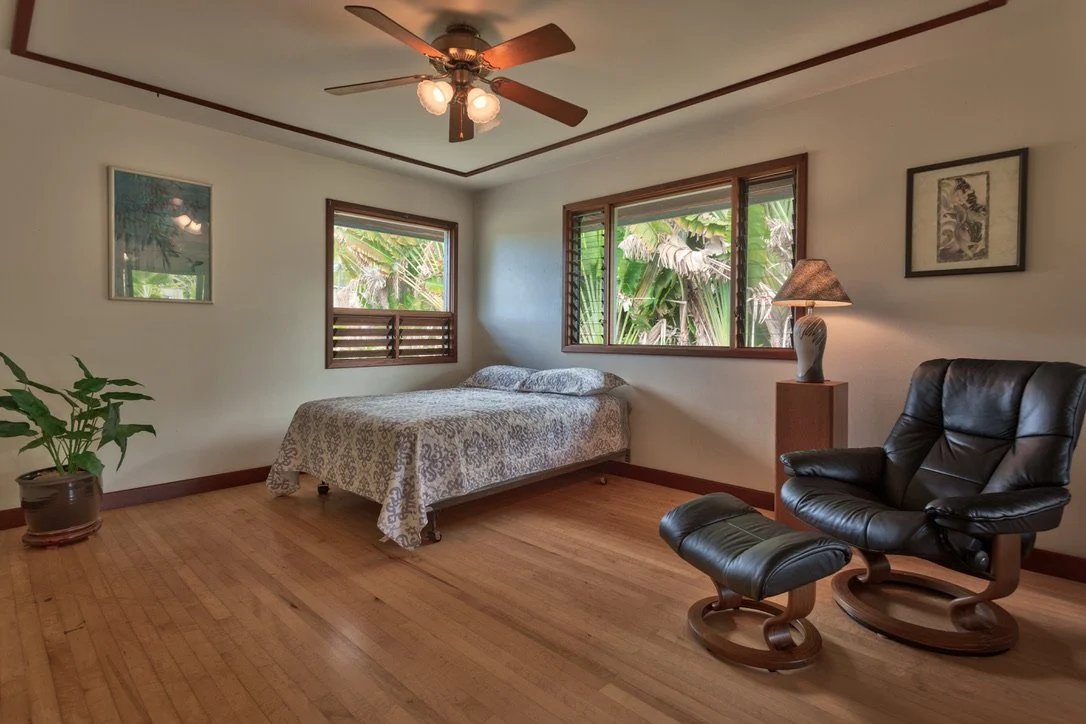A bedroom with a bed, a potted plant, a leather chair with matching footstool, a nightstand with a lamp, and three picture frames on the walls. Large windows show greenery outside.