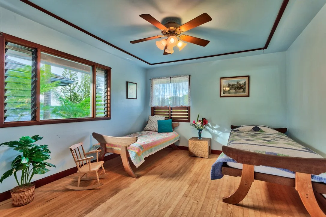 A bedroom with light blue walls and wooden floors, featuring two wooden beds with striped and colorful bedding, a ceiling fan with lights, a window with blinds and lace curtains, a small nightstand with a vase of flowers, a leafy green plant in a basket, and two framed pictures on the walls.