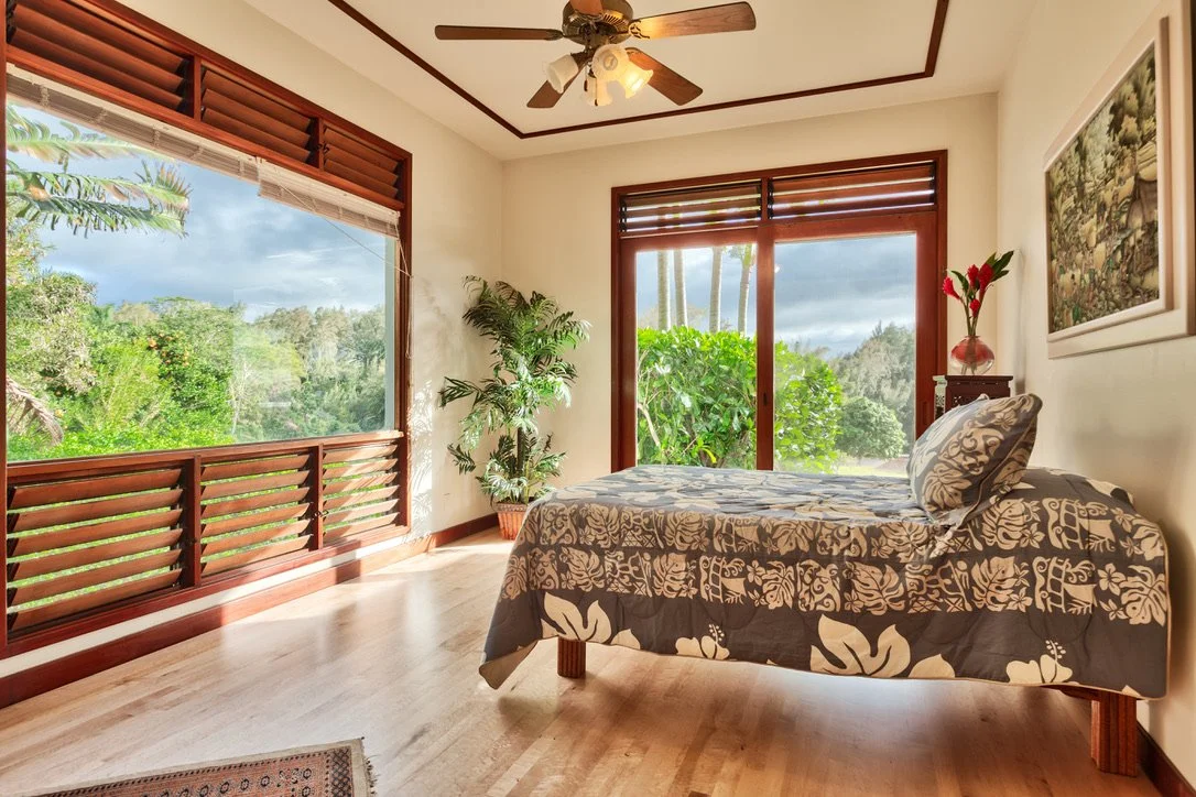 A bedroom with a bed, potted plant, framed picture, and large windows showing lush green trees outside under a cloudy sky.