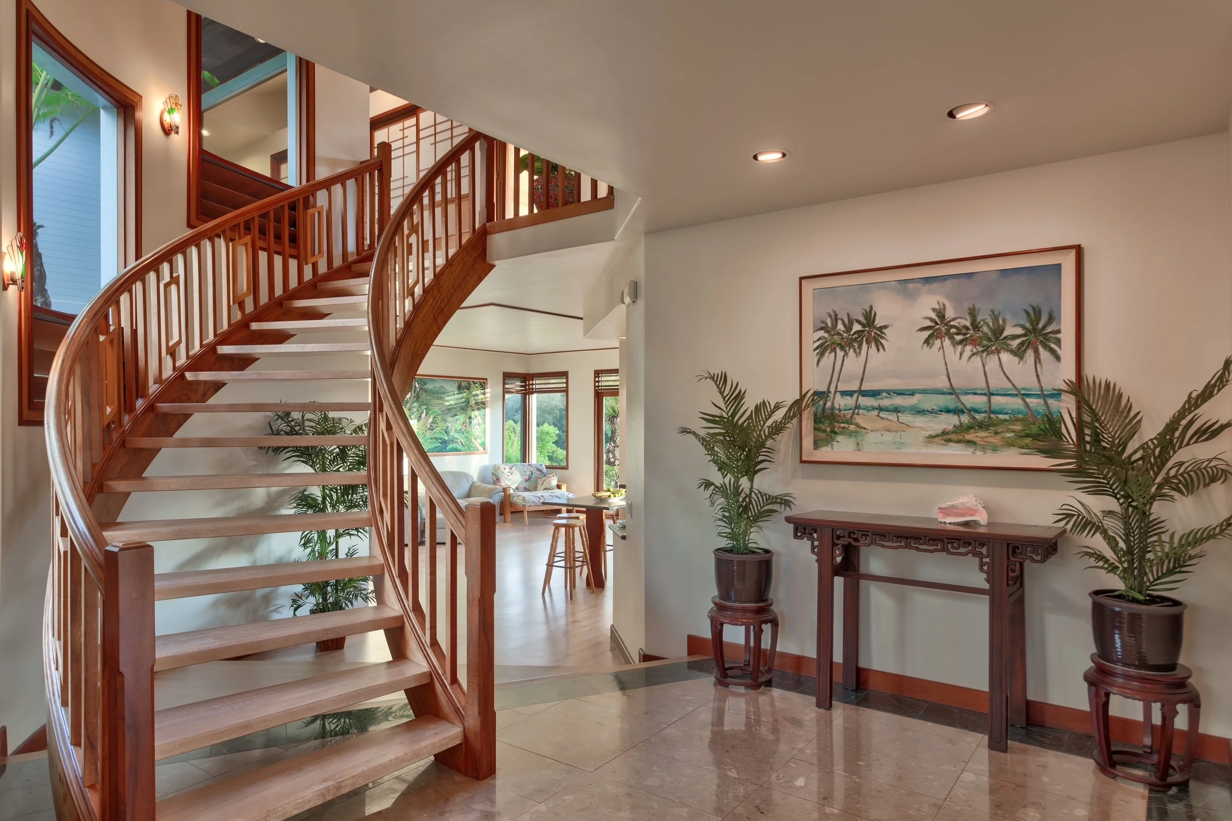 Interior view of a home with a curved wooden staircase, tropical-themed artwork on the wall, potted plants, and large windows showing greenery outside.