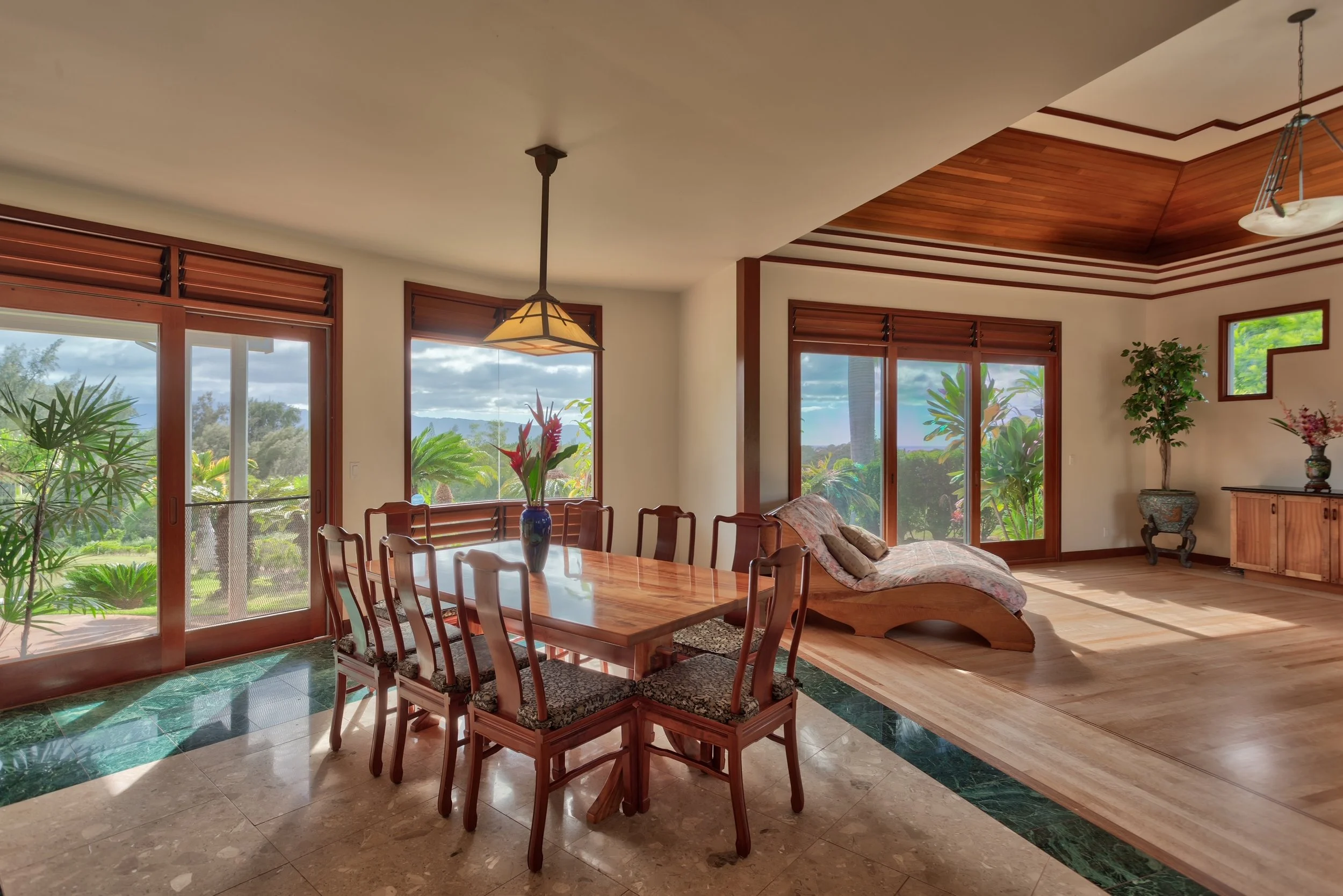 Living room with large windows, wooden dining table with chairs, a floral vase, and plant decorations, featuring a wooden ceiling and a mix of tile and parquet flooring.