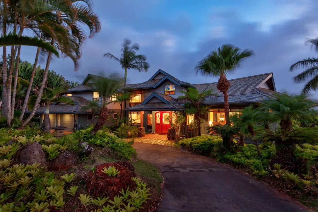 A large house with a driveway surrounded by tropical plants and palm trees, illuminated with warm indoor lighting during dusk.