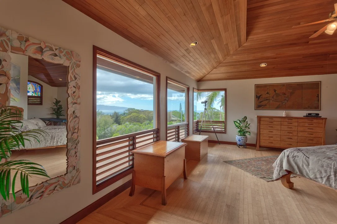 Bedroom with large windows showing an ocean and tropical view, wooden ceiling, dresser, potted plant, and bed with white bedding.