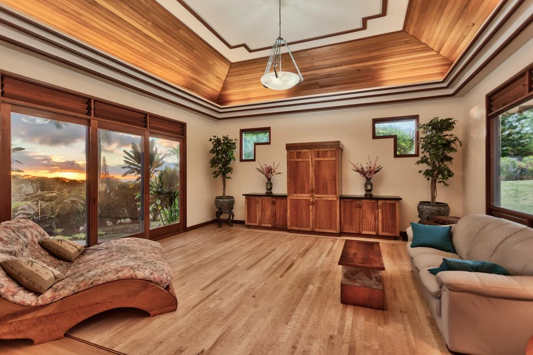 Living room with wooden flooring, large windows showing sunset, potted plants, wooden cabinets, and sofas.