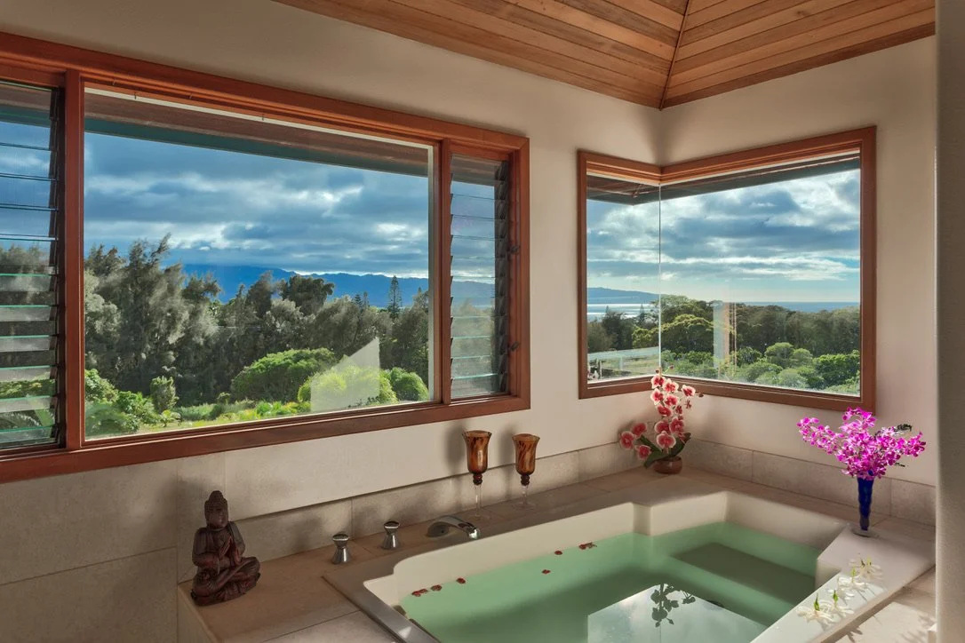 Bathroom with a soaking tub filled with water, decorated with flower petals, and overlooking a lush green landscape through large windows, with a wooden ceiling and decorative vases and flowers around the tub.
