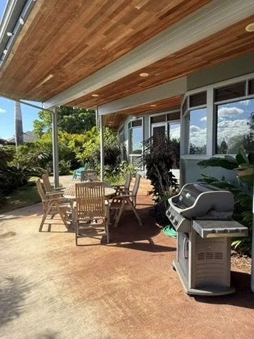 Outdoor patio area with wooden chairs and a table, barbecue grill, and lush greenery in the background under a wooden overhang.