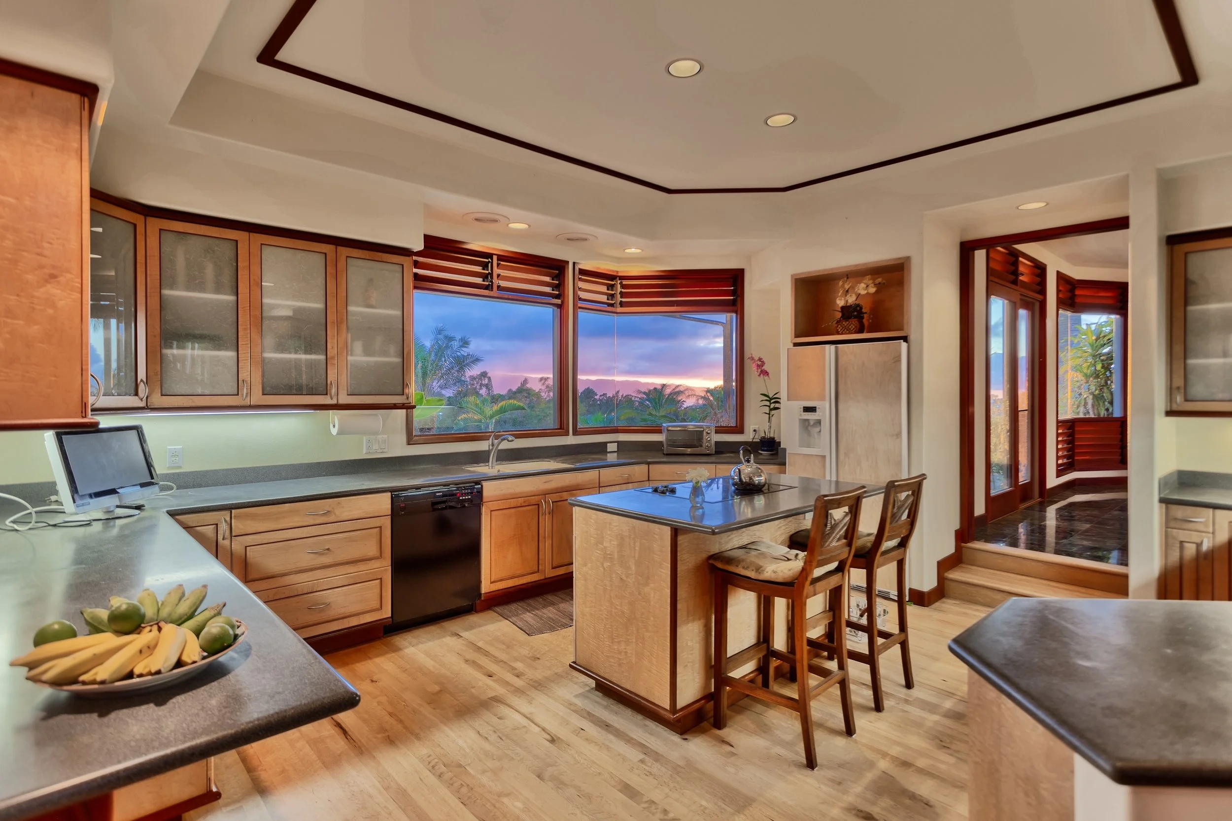 A spacious kitchen with wooden cabinets, large windows showing a sunset and trees outside, a kitchen island with bar stools, a bowl of bananas and limes, and various small appliances on the counters.