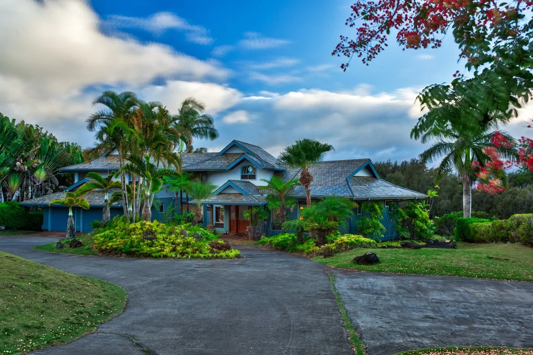 A large tropical-style house with a curved driveway, surrounded by palm trees and lush greenery, under a partly cloudy sky.