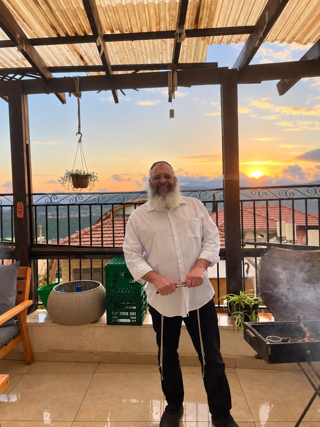 A man with a beard and glasses standing on a balcony at sunset, holding grill tongs, with a barbecue grill nearby and potted plants around.