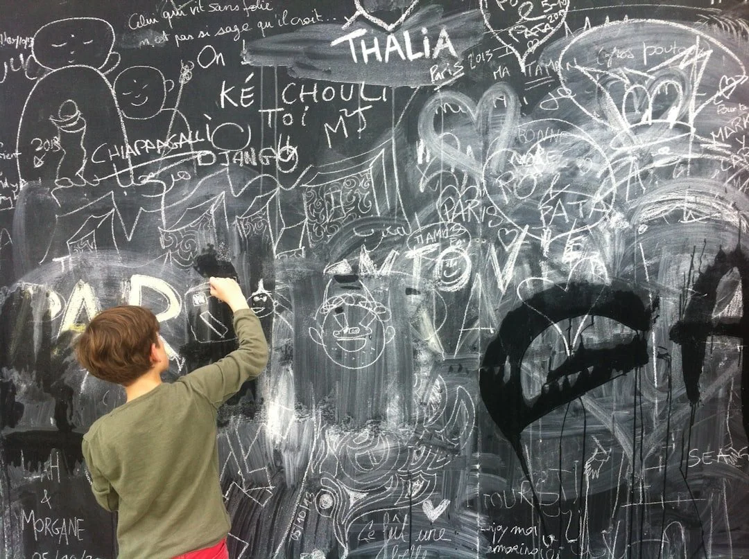A young boy with brown hair in a green sweater is drawing and writing on a large black chalkboard filled with doodles, writings, and graffiti in white chalk, including hearts, faces, words, and names.