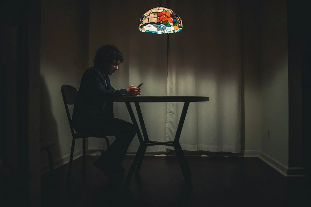 A man sitting at a table in a dimly lit room, looking at his phone, with a colorful stained glass lamp hanging above him.