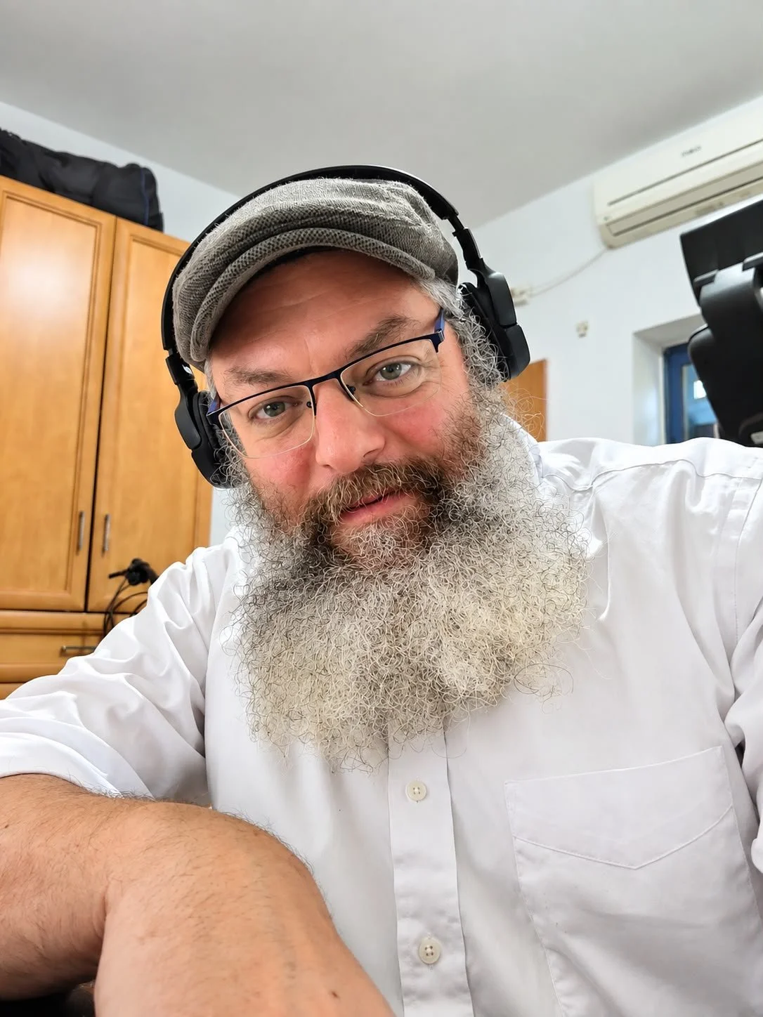 A man with glasses, a gray beard, and wearing headphones and a cap, takes a selfie indoors, with a wooden cabinet and window in the background.