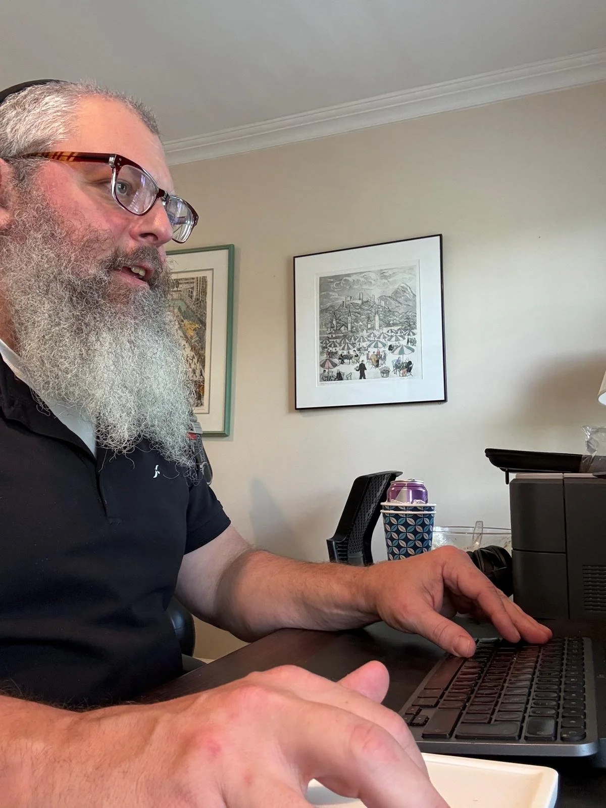 An older man with gray hair, glasses, and a beard sitting at a desk, working on a laptop. Behind him, there are framed pictures on the wall, one of which depicts a beach scene with umbrellas and people. On the desk, there is a purple beverage can and a patterned cup holder.