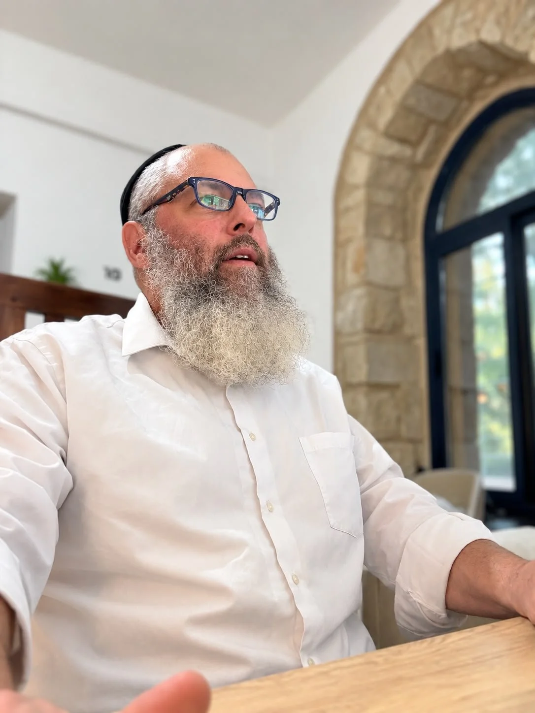 A man with gray hair and beard wearing glasses and a white shirt, sitting indoors near a large arched window with a brick surround, during daytime.