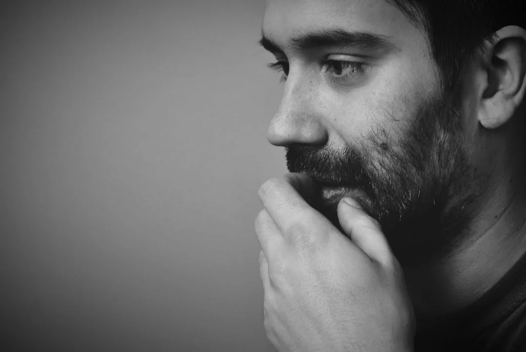 A black and white photo of a man with a beard, seen in profile, touching his lips with his fingers, looking thoughtful.