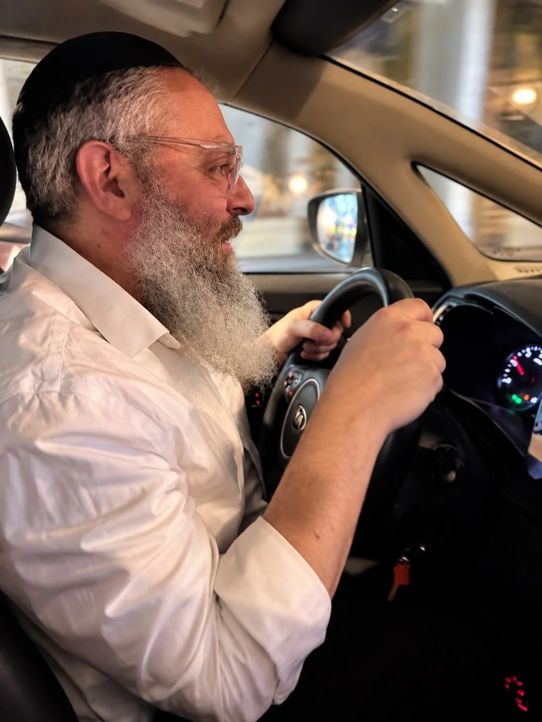 An elderly man with a white beard, wearing glasses and a black kippah, driving a car while holding the steering wheel with both hands.