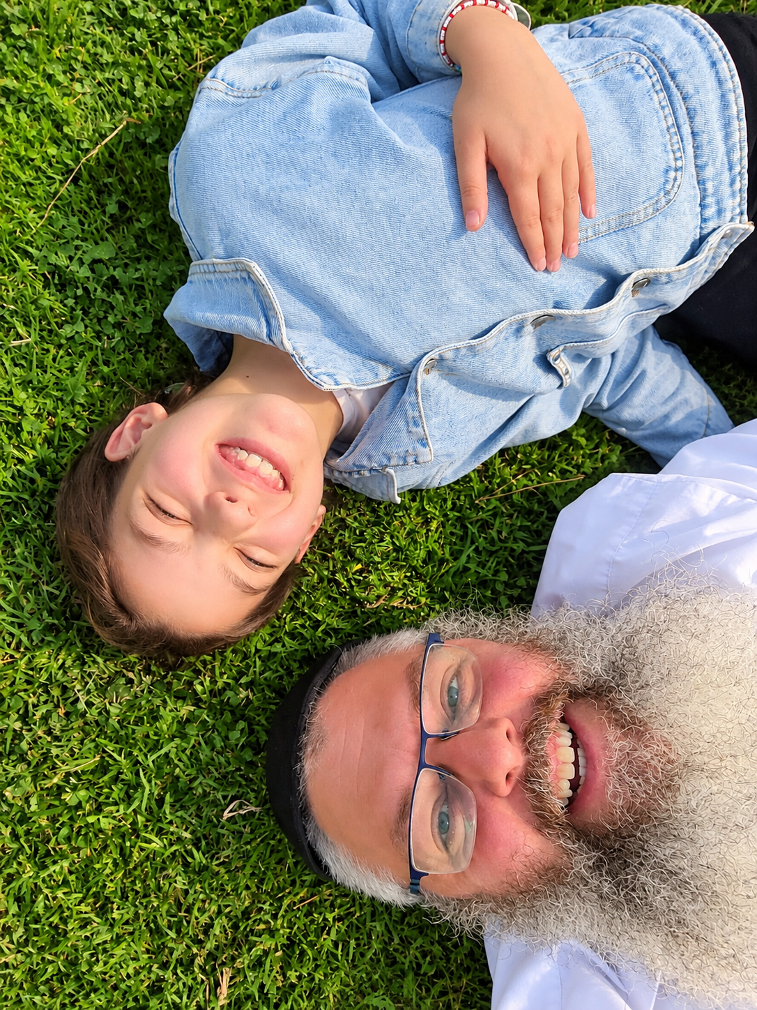 A young boy and an older man with a beard and glasses lying on green grass, smiling and looking up.