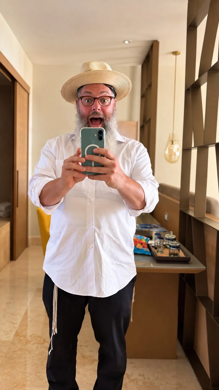A man taking a selfie in a mirror, wearing a straw hat, glasses, a white shirt, and black pants, with a surprised or excited expression.