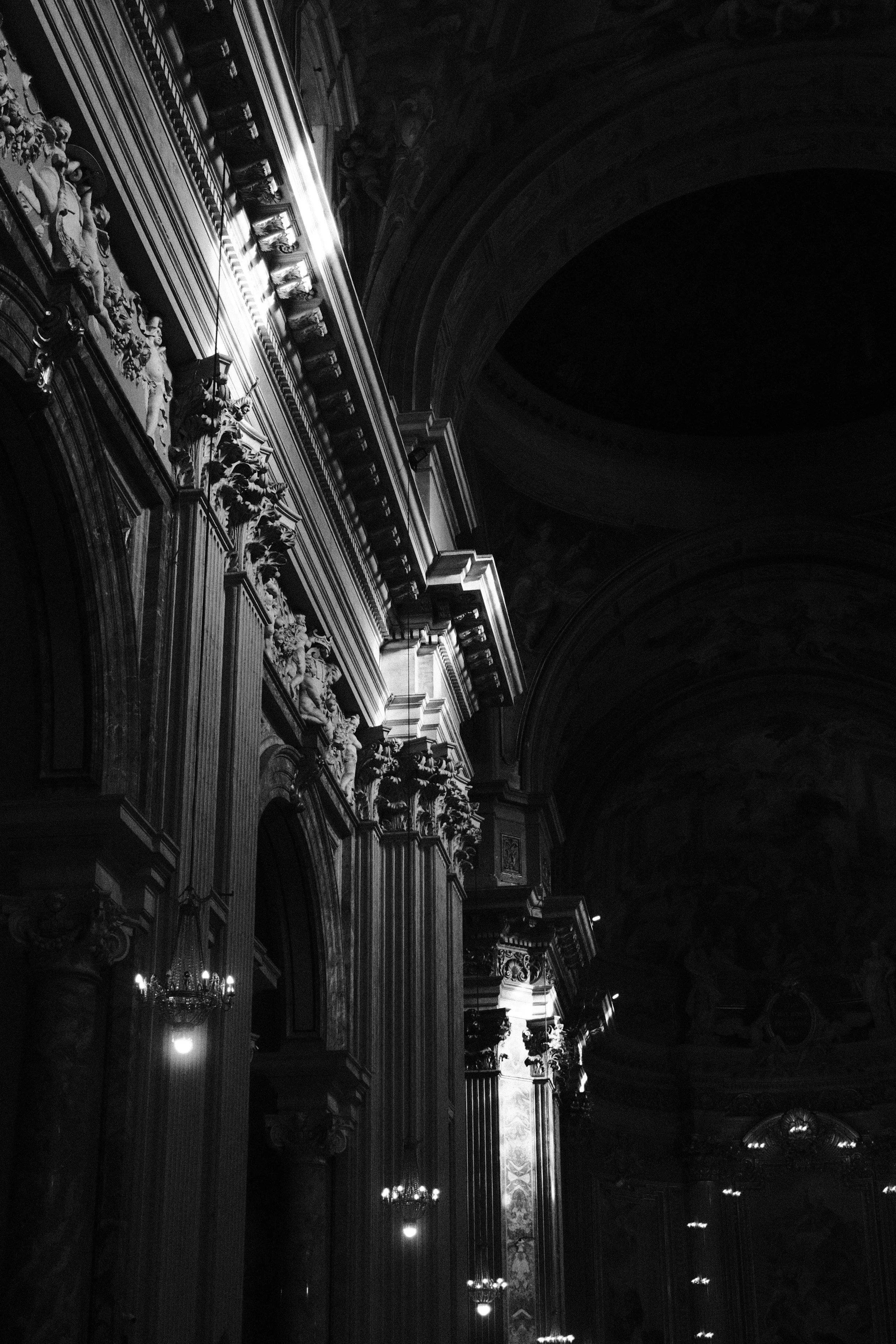 Interior view of a grand, ornate hall with detailed carvings, chandeliers, and high arched ceilings, illuminated by wall sconces.
