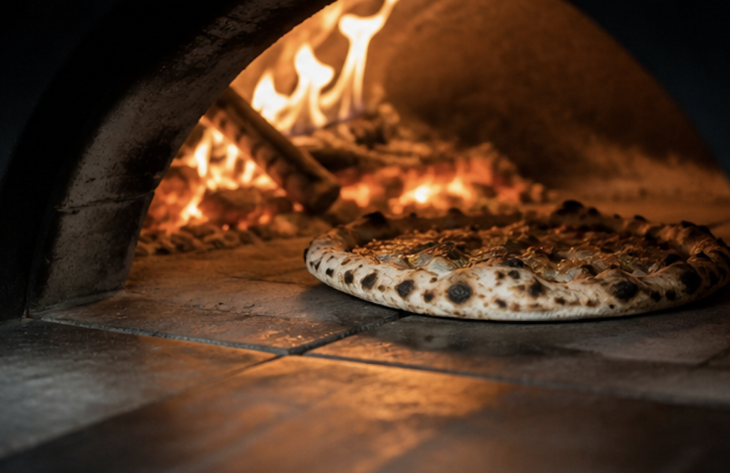 Pizza baking inside a wood-fired pizza oven with flames visible.